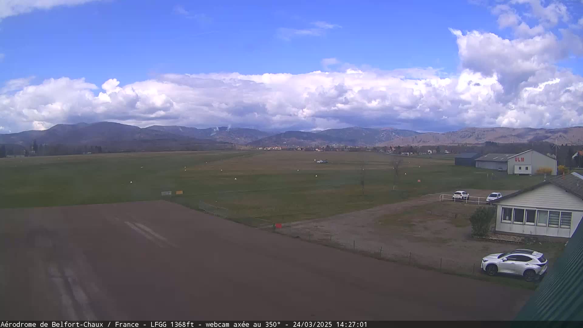A mostly sunny airfield with a few puffy clouds in the sky, shows a runway, some buildings, cars, and distant mountains.