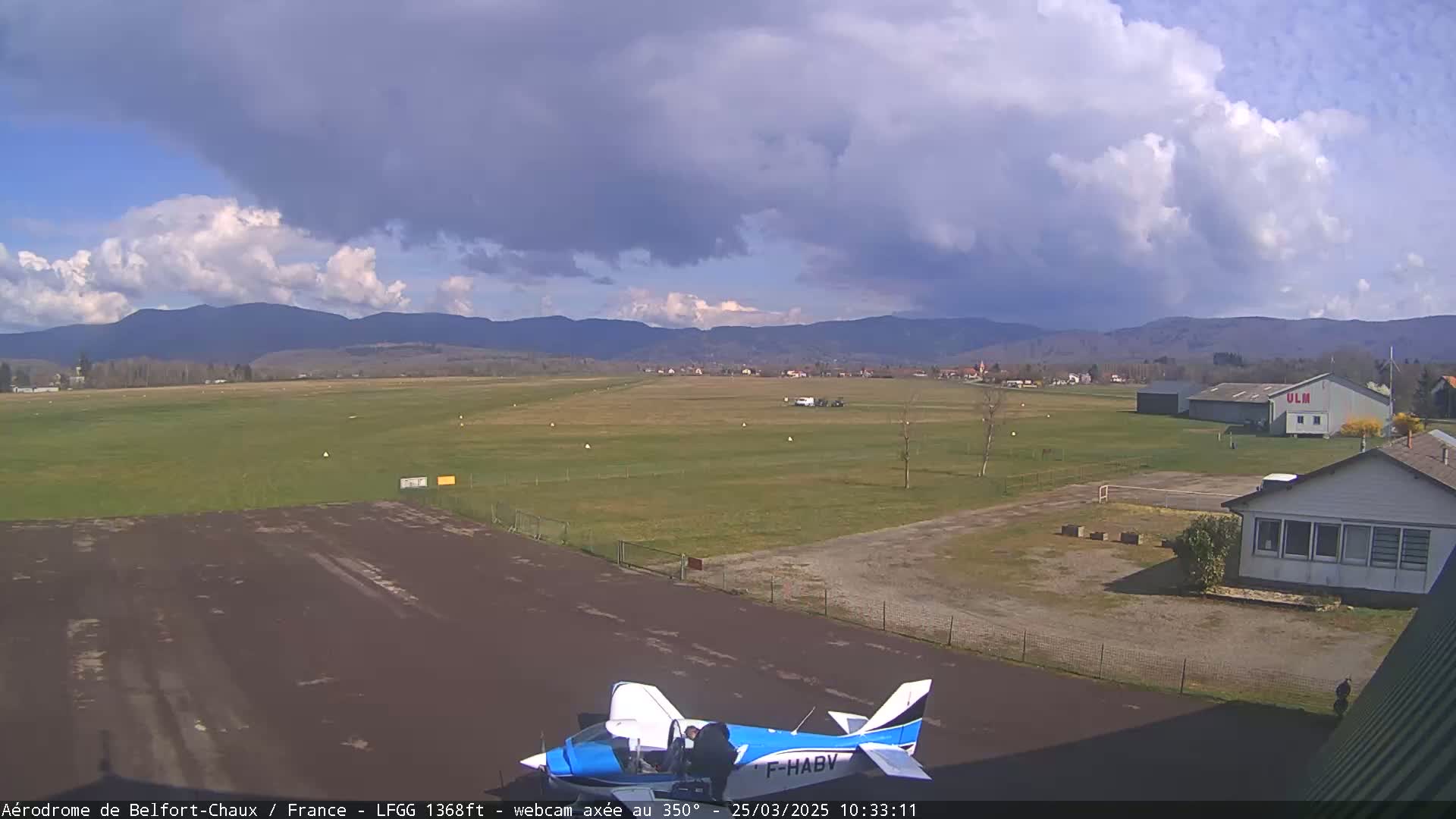 A small airplane is parked on a tarmac at an airfield under partly cloudy skies with mountains in the distance.