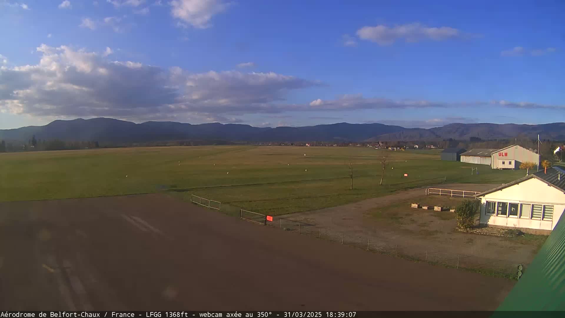 A grassy airfield with a small building and hangar is visible under a partly cloudy sky, with mountains in the background.