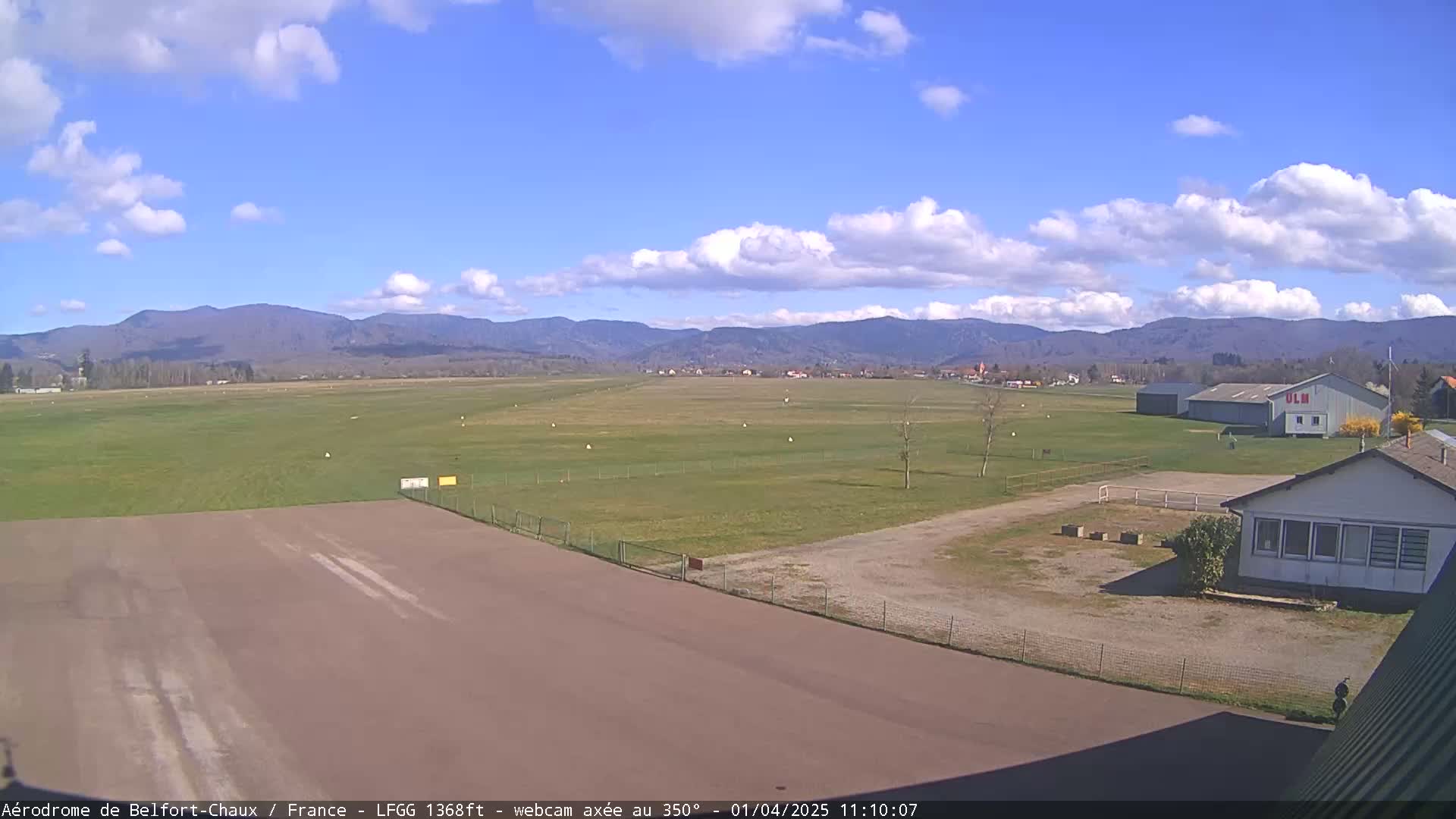 A grassy airfield with a runway and small buildings is visible under a partly cloudy blue sky, with mountains in the distance.