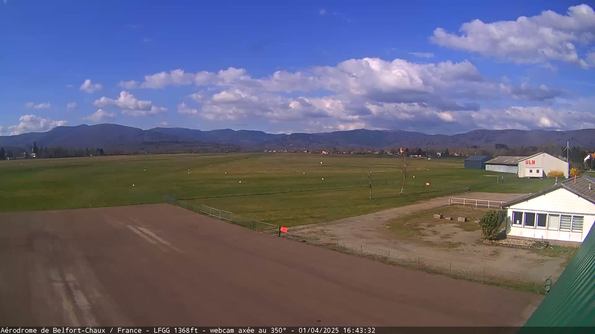 A mostly sunny day reveals a grassy airfield with a runway, hangars, and a small building, backed by distant mountains under a partly cloudy sky.
