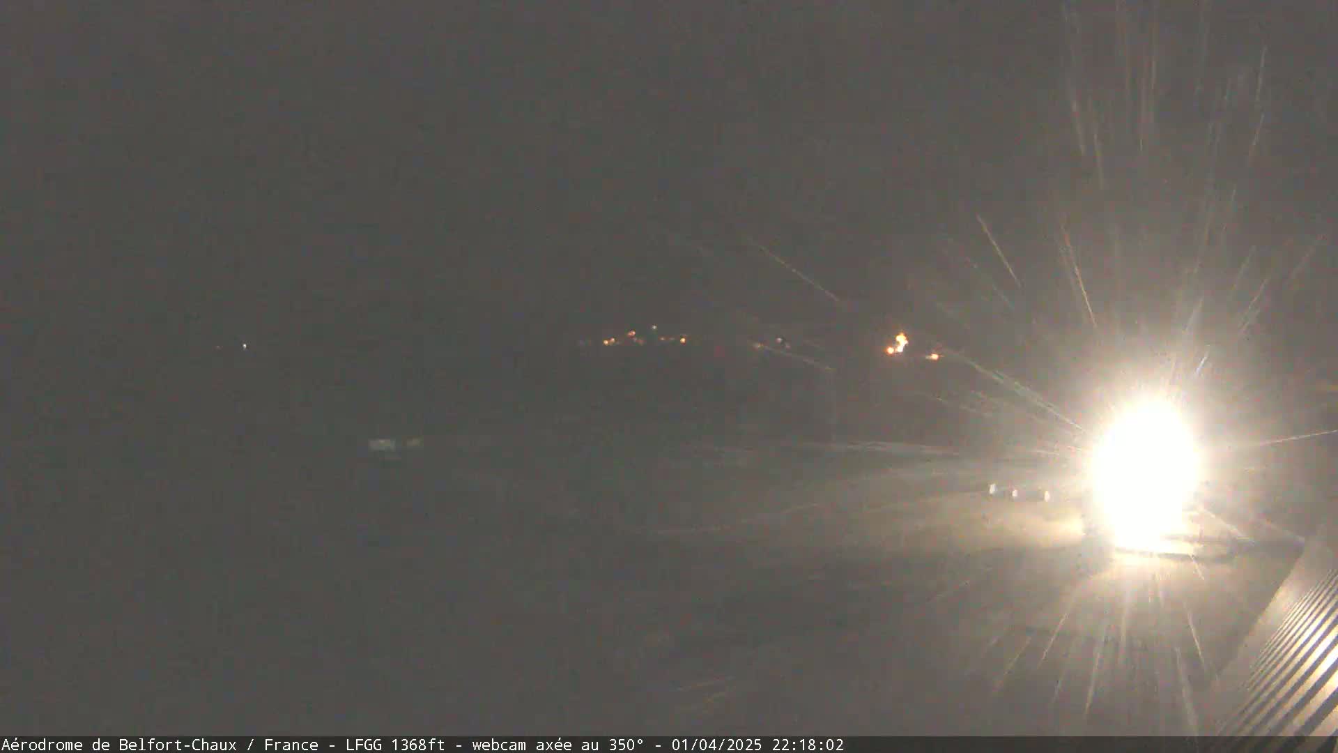 A bright light shines intensely in the foreground of a dark, snowy nighttime scene, with a few distant lights visible in the background.