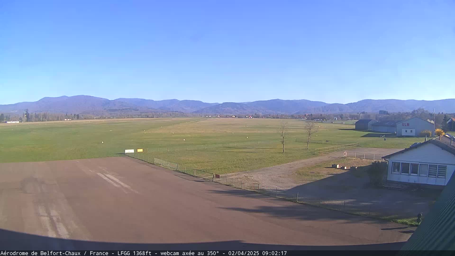 A small airfield with a runway and a few buildings is situated before a wide expanse of grassy field and distant mountains under a clear blue sky.