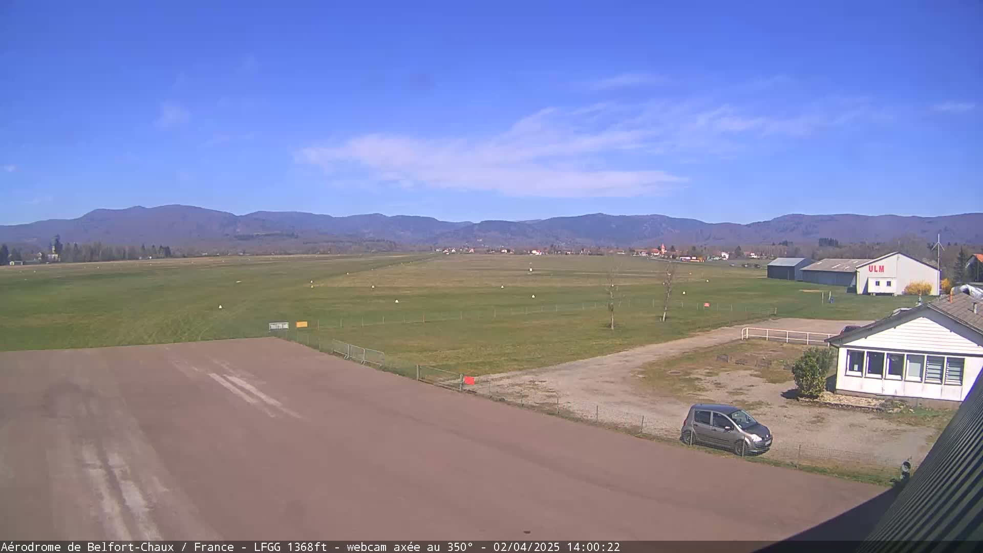 A small airfield with a runway, grassy areas, a hangar, and a single car is visible under a clear blue sky.