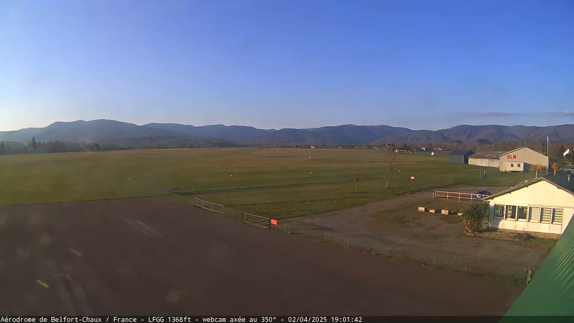 A flat, grassy airfield with a small building and hangar is visible, under a clear blue sky, with distant mountains in the background.