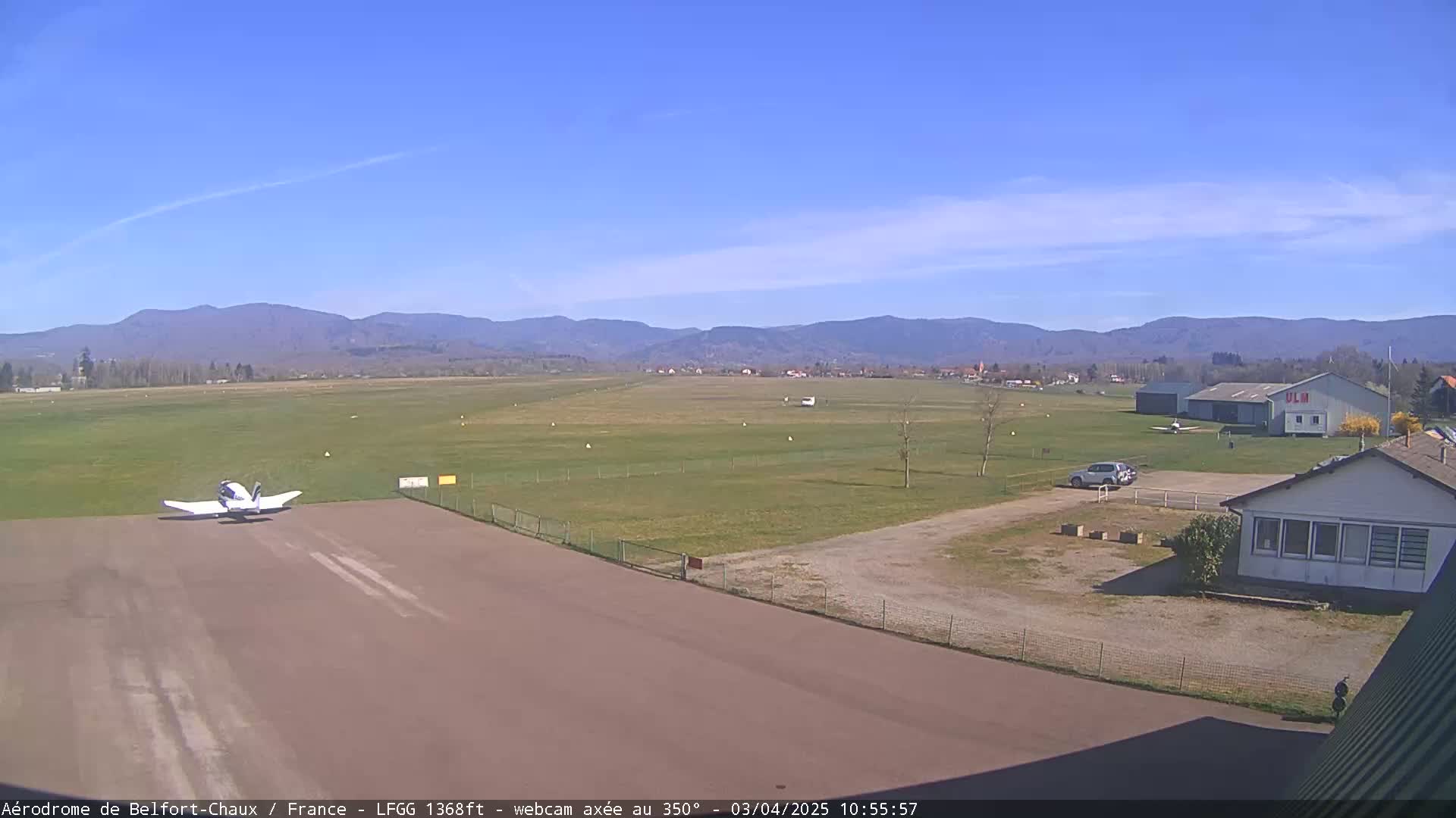 A small airplane taxis on a runway at an airfield under a clear blue sky, with mountains visible in the distance.