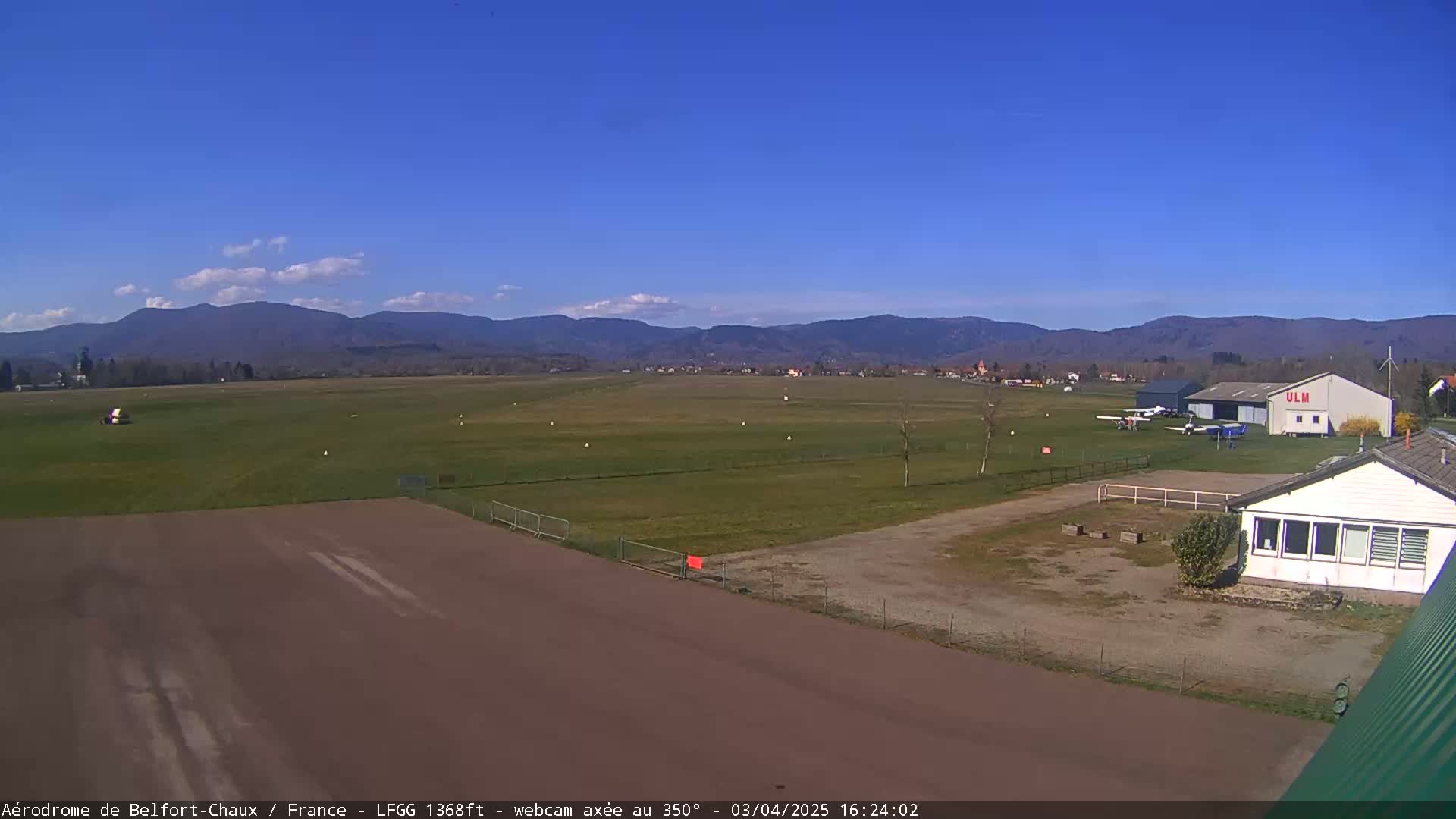 A small airfield with several small aircraft is shown, under a mostly clear blue sky with some scattered clouds, in front of a range of distant mountains.