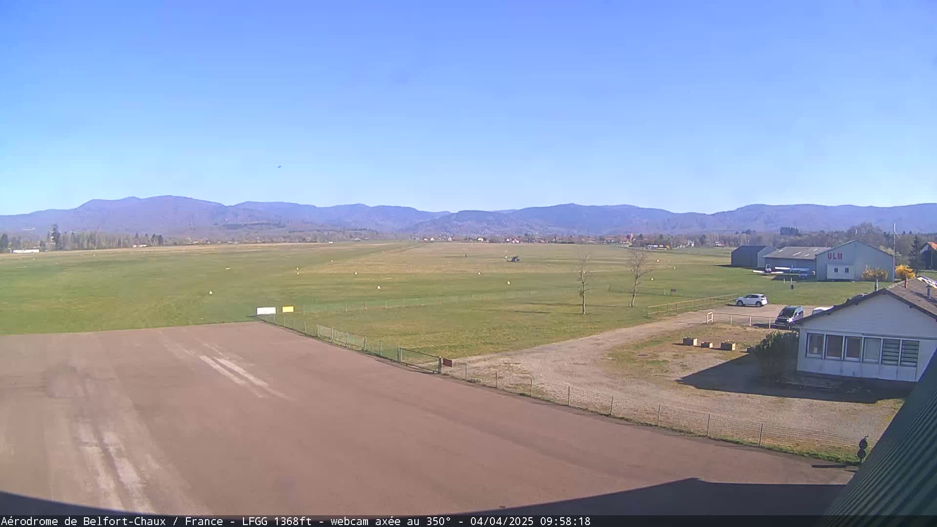 A small airfield with a runway, several buildings, and a few cars is visible under a clear blue sky, with mountains in the distance.