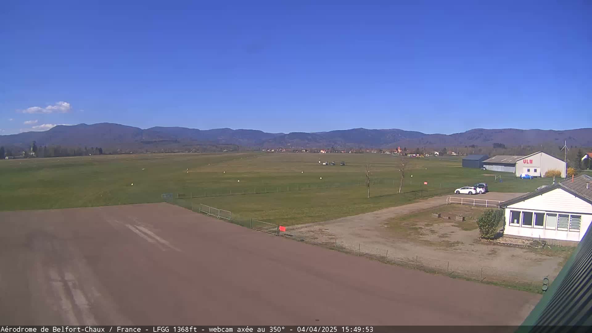 A small airfield with a hangar and a small building is visible, with a clear blue sky and distant mountains in the background.