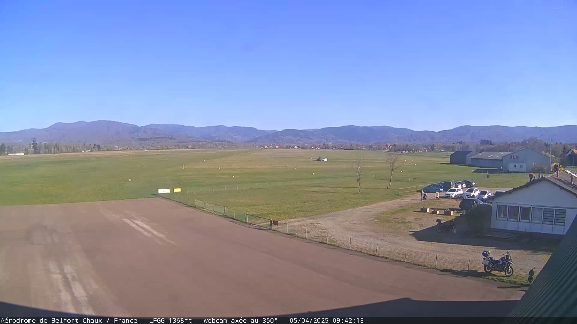 A grassy airfield with a runway, several parked cars and a building, is situated in front of a mountain range under a clear blue sky.