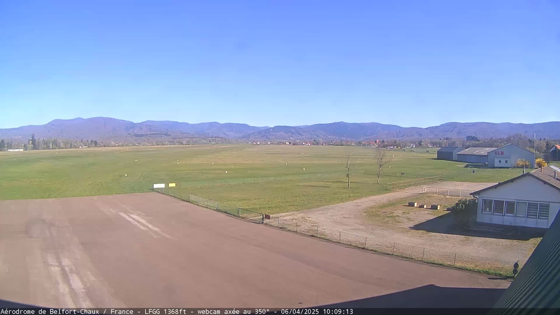A small airfield with a runway and several buildings is seen under a clear blue sky, with distant mountains in the background.