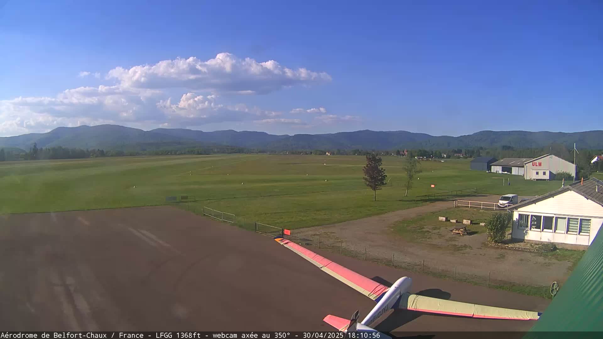 A glider's wing is visible in the foreground of an airfield under a partly cloudy blue sky, with distant mountains in the background.