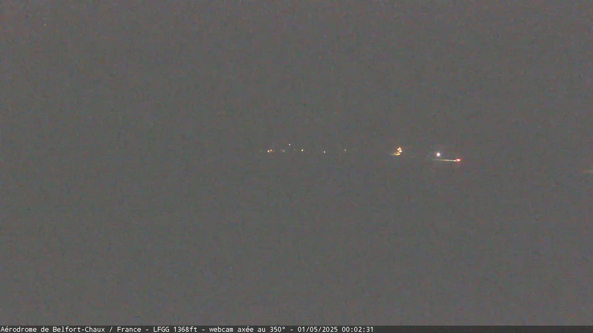 A nighttime long shot of a dark landscape with a few faint lights in the distance under a dark sky.