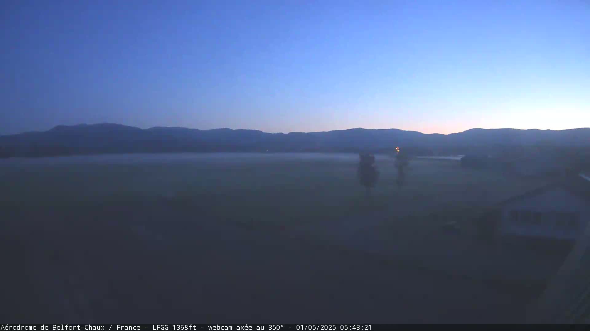 A foggy, pre-dawn scene shows a flat expanse of land with distant mountains under a clear, dark-blue sky.