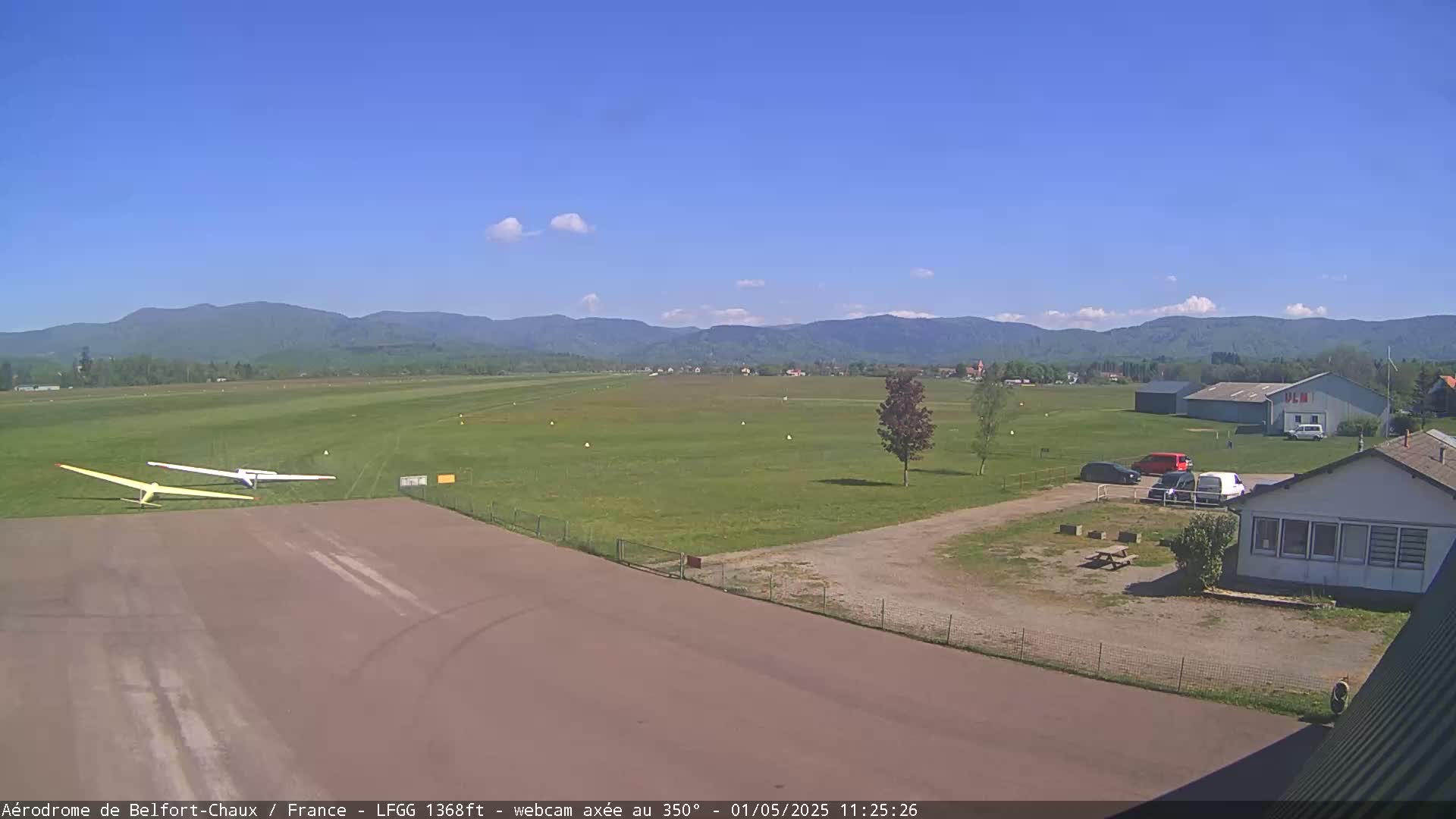 Two gliders are parked on a tarmac next to a grassy airfield, with a small building and several cars nearby, all under a clear blue sky with a few clouds and distant mountains in the background.