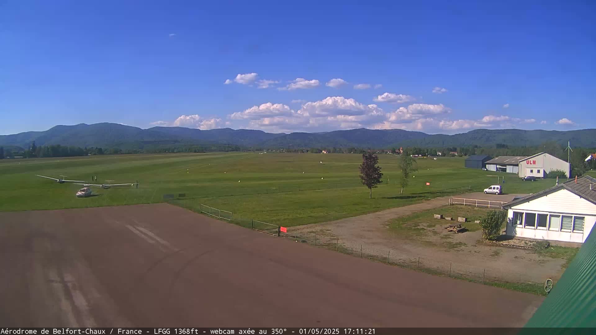 A small airfield with gliders and a car, situated in front of a building, under a partly cloudy sky and backed by a mountain range.