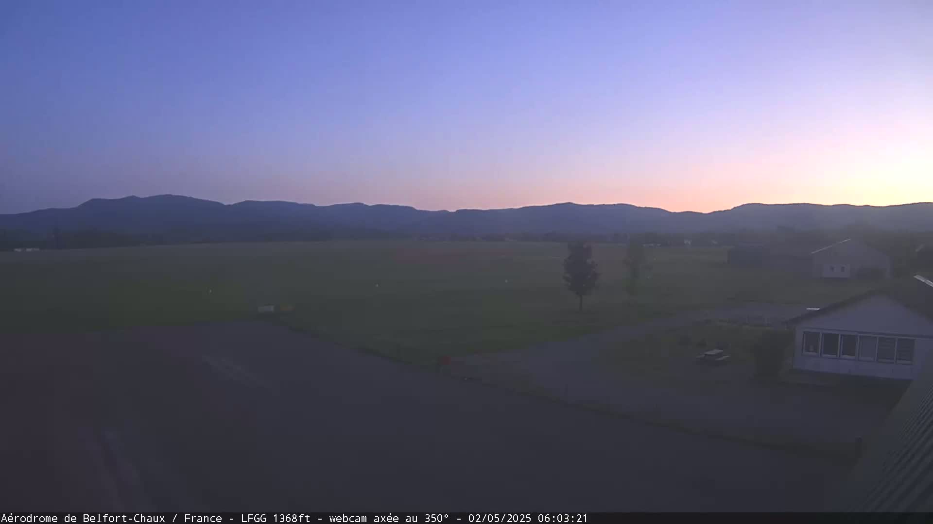 A twilight view shows a flat green field, a few buildings, and distant mountains under a clear, dusky sky.