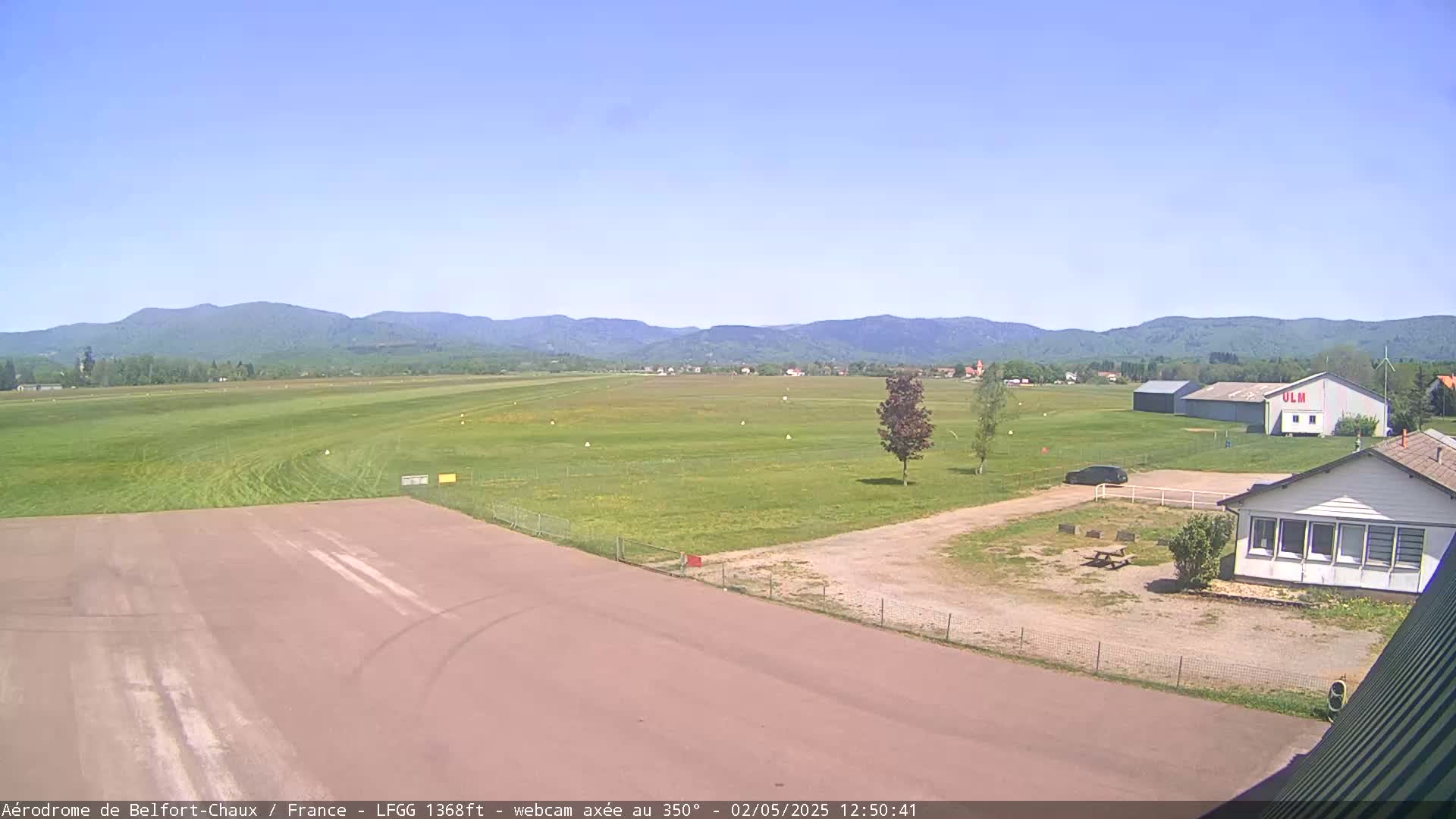 A small airfield with a runway, several buildings, and a few cars is situated in a grassy field, with mountains visible in the background under a clear blue sky.