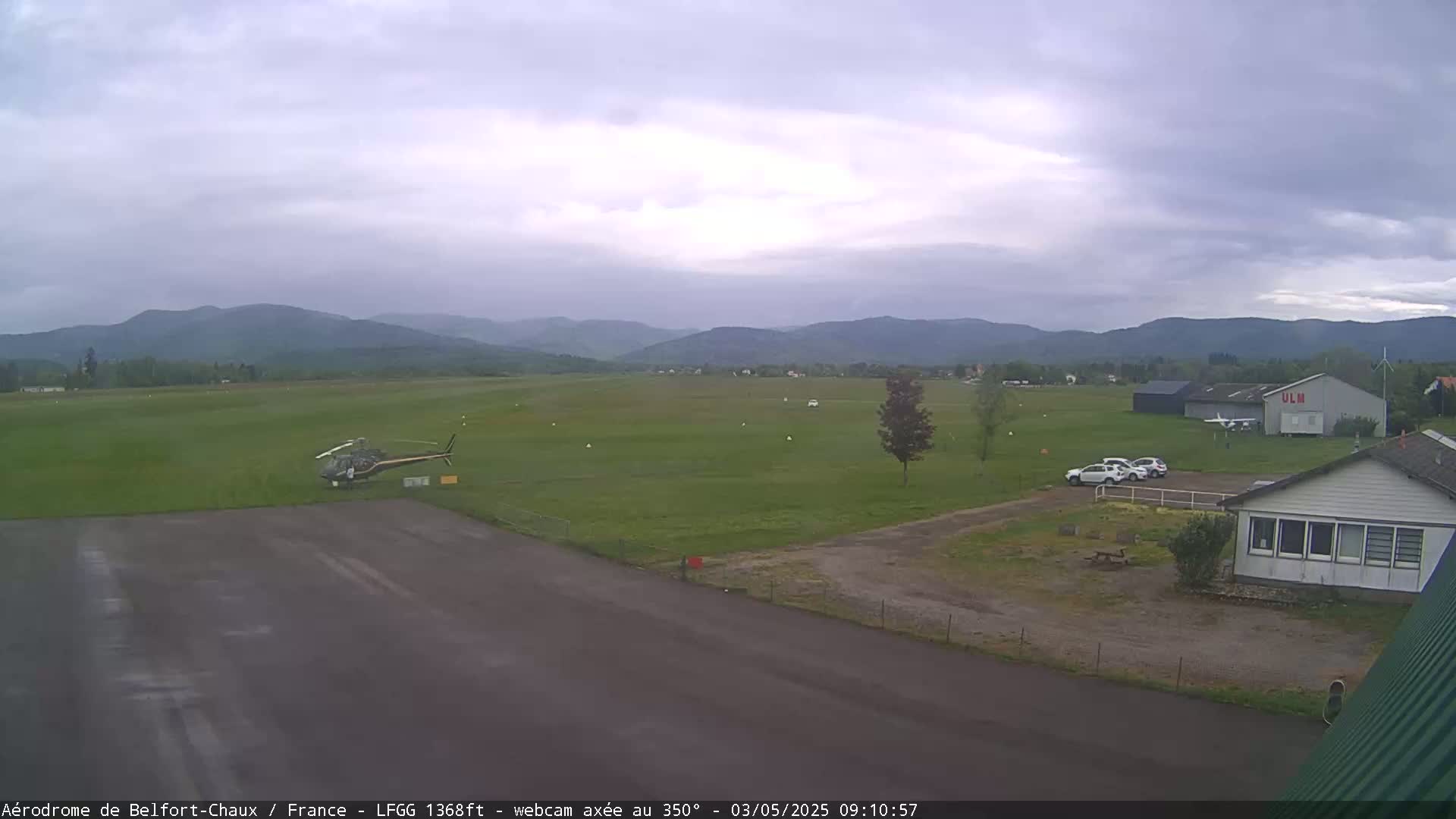 A helicopter sits on a tarmac at a small airfield, with overcast skies and distant mountains in the background.