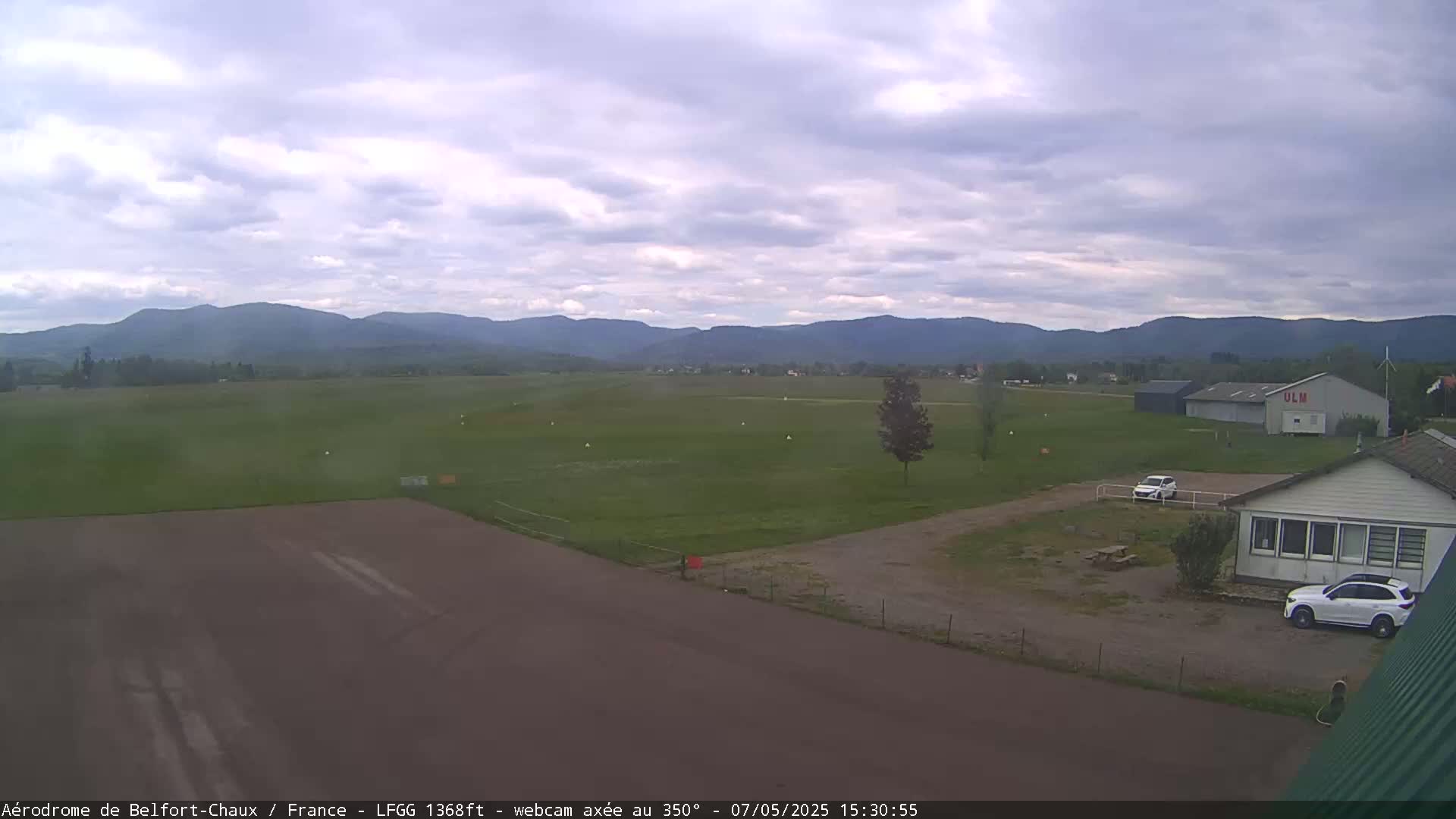 A mostly cloudy day reveals a grassy airfield with several buildings, two cars, and distant mountains.