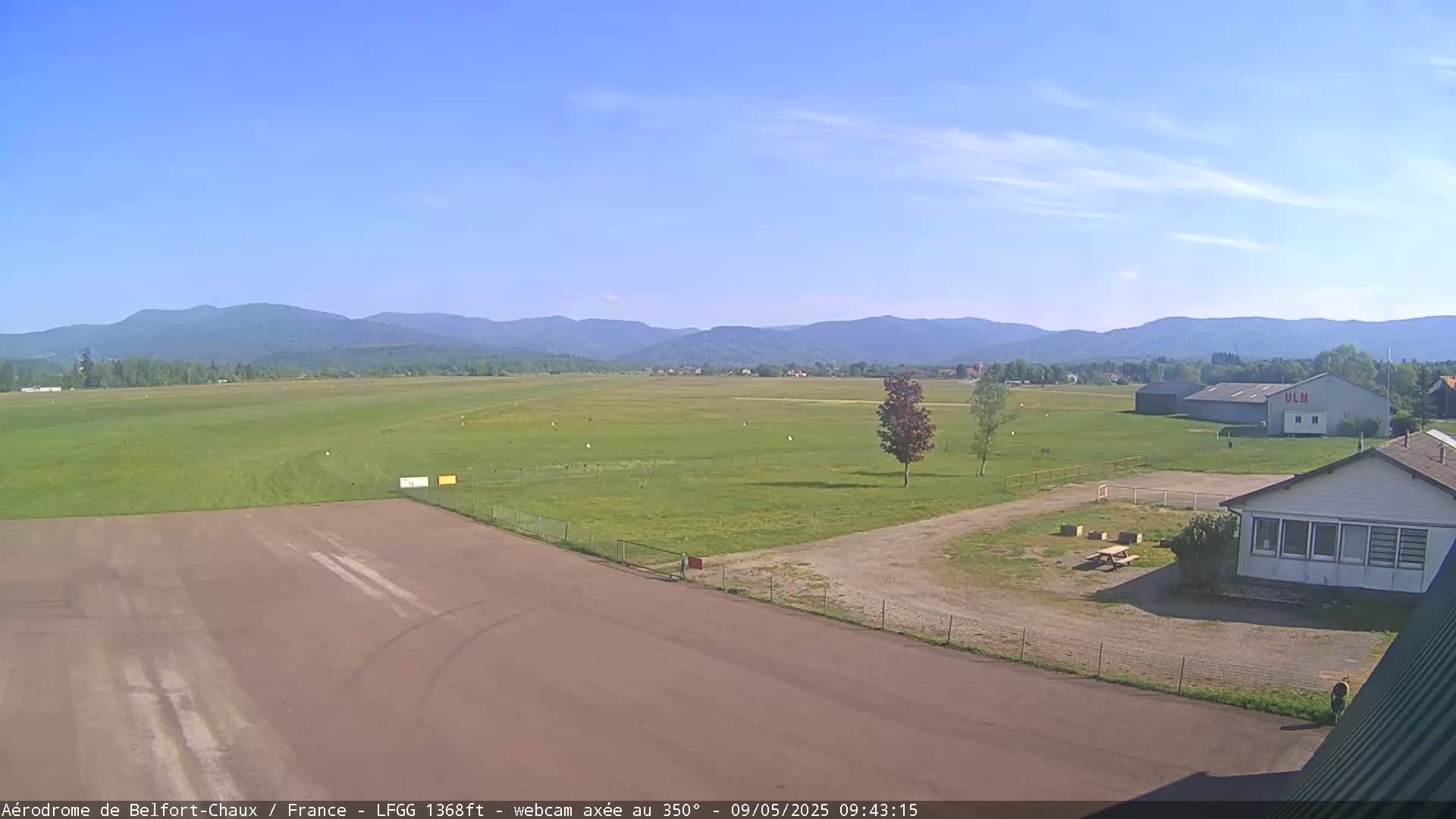 A grassy airfield with a runway and buildings is visible under a clear blue sky, with mountains in the background.