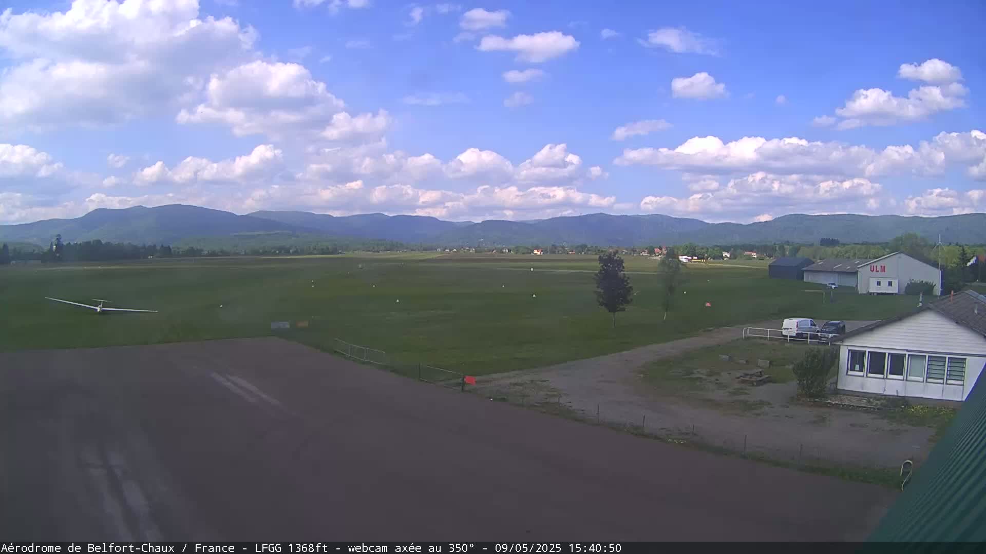 A glider sits on a grassy airfield under a partly cloudy sky, with distant mountains visible in the background.