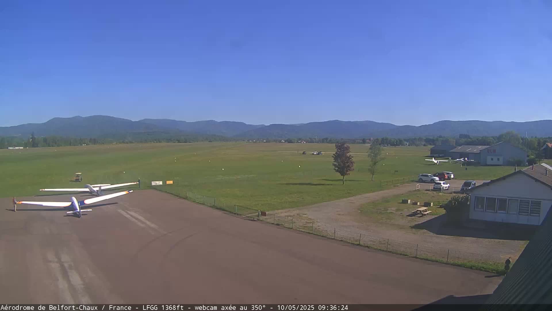 Three gliders are parked on a tarmac at a small airfield on a clear, sunny day, with distant mountains visible in the background.