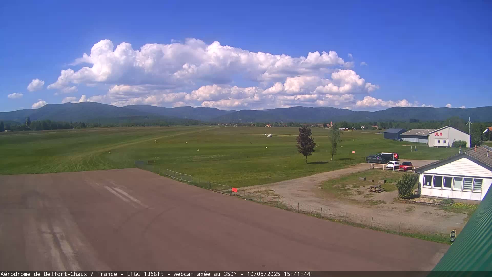 A small airfield with a few parked cars and buildings is visible under a partly cloudy sky, with mountains in the background.