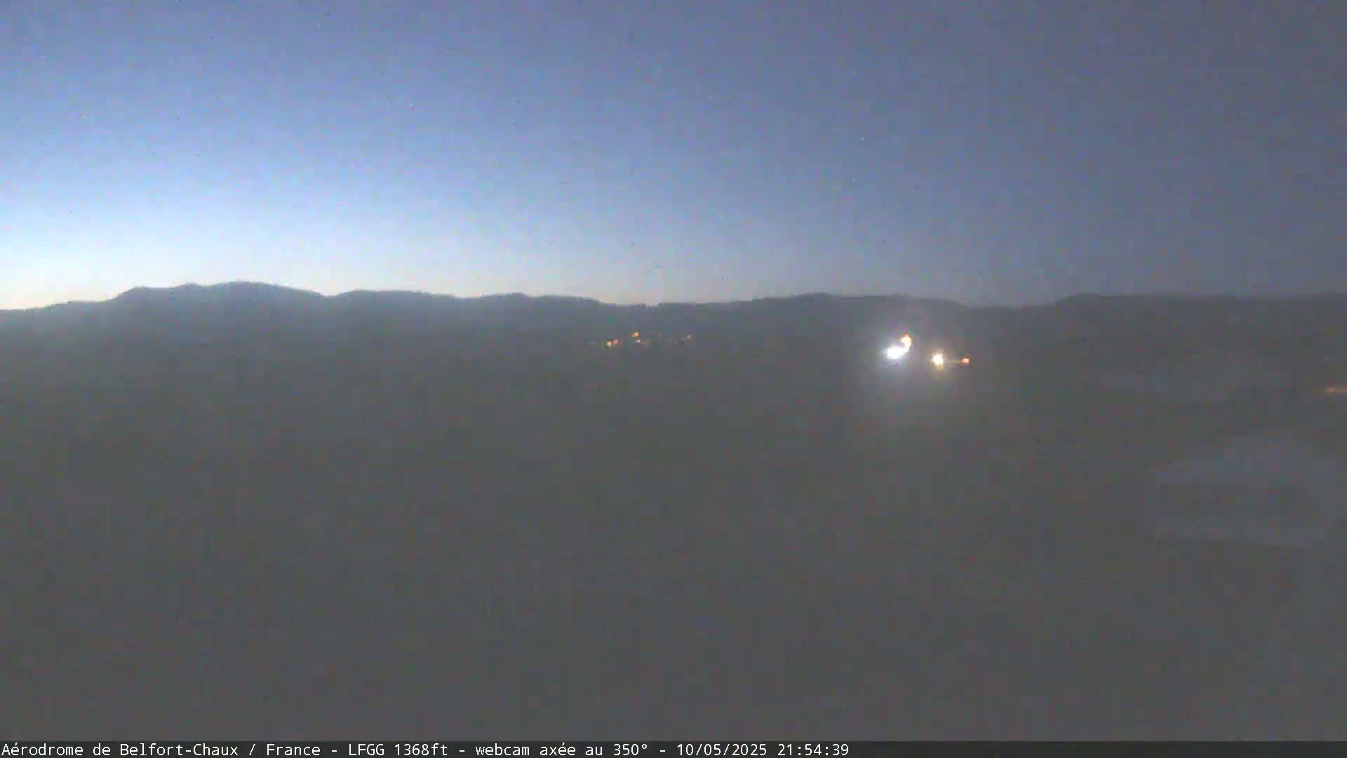A hazy, dark twilight scene shows distant hills and a few faint lights in the foreground.