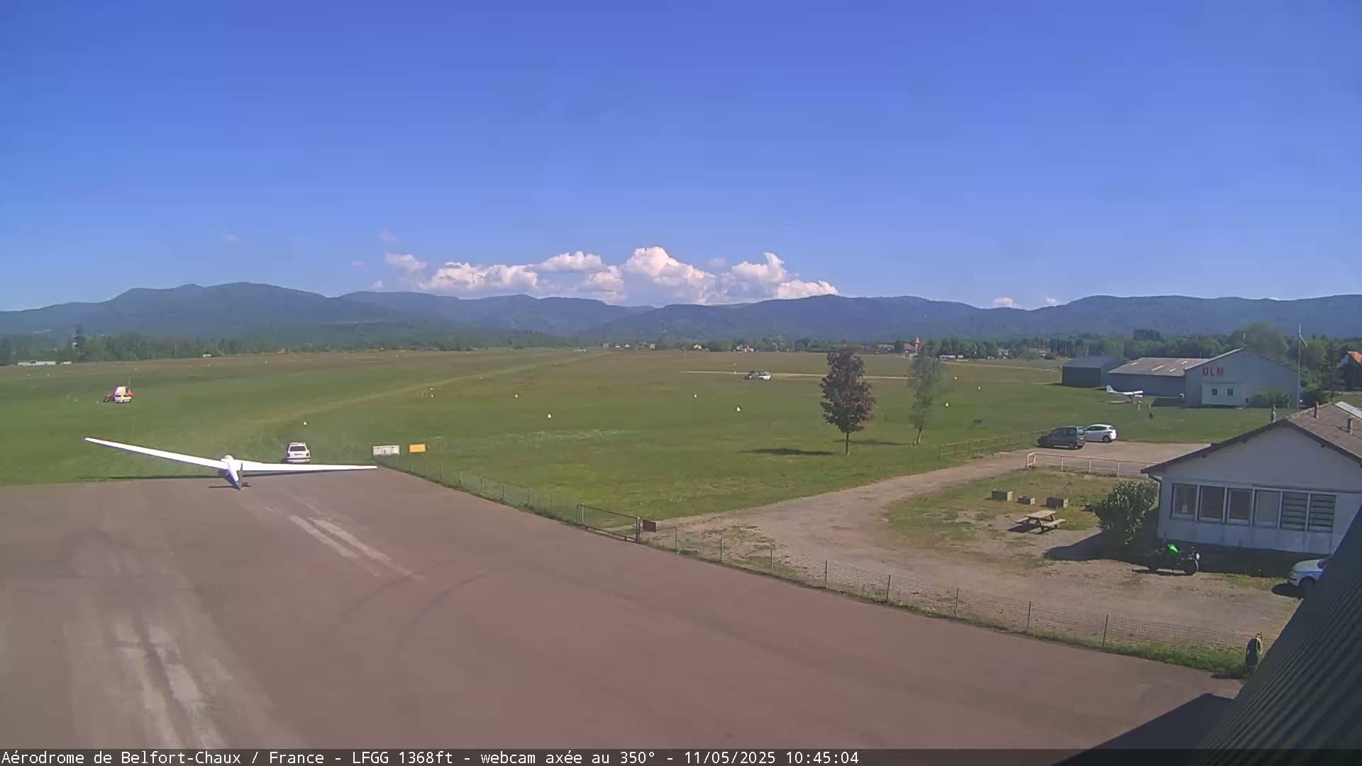 A glider sits on a runway at a small airfield on a sunny day with partly cloudy skies and mountains in the background.