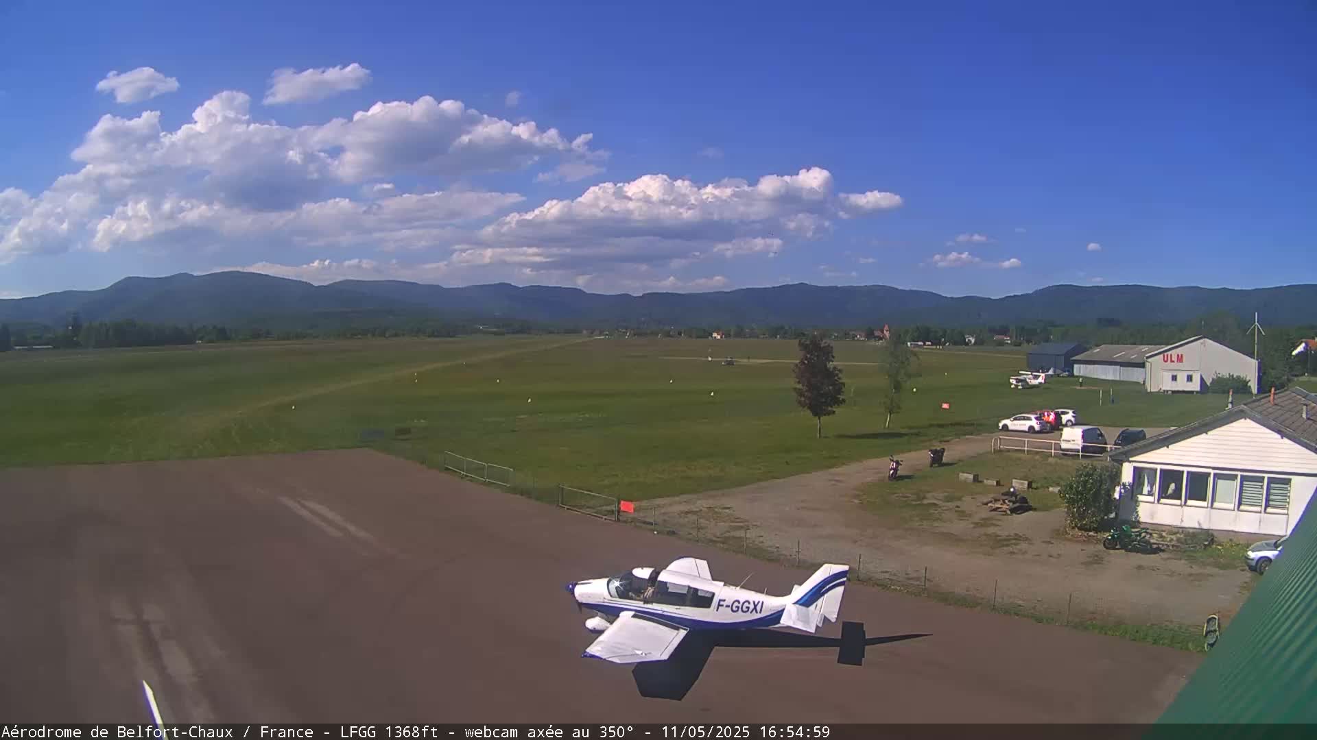 A small airplane sits on a tarmac at an airfield under a partly cloudy blue sky, with mountains visible in the distance.