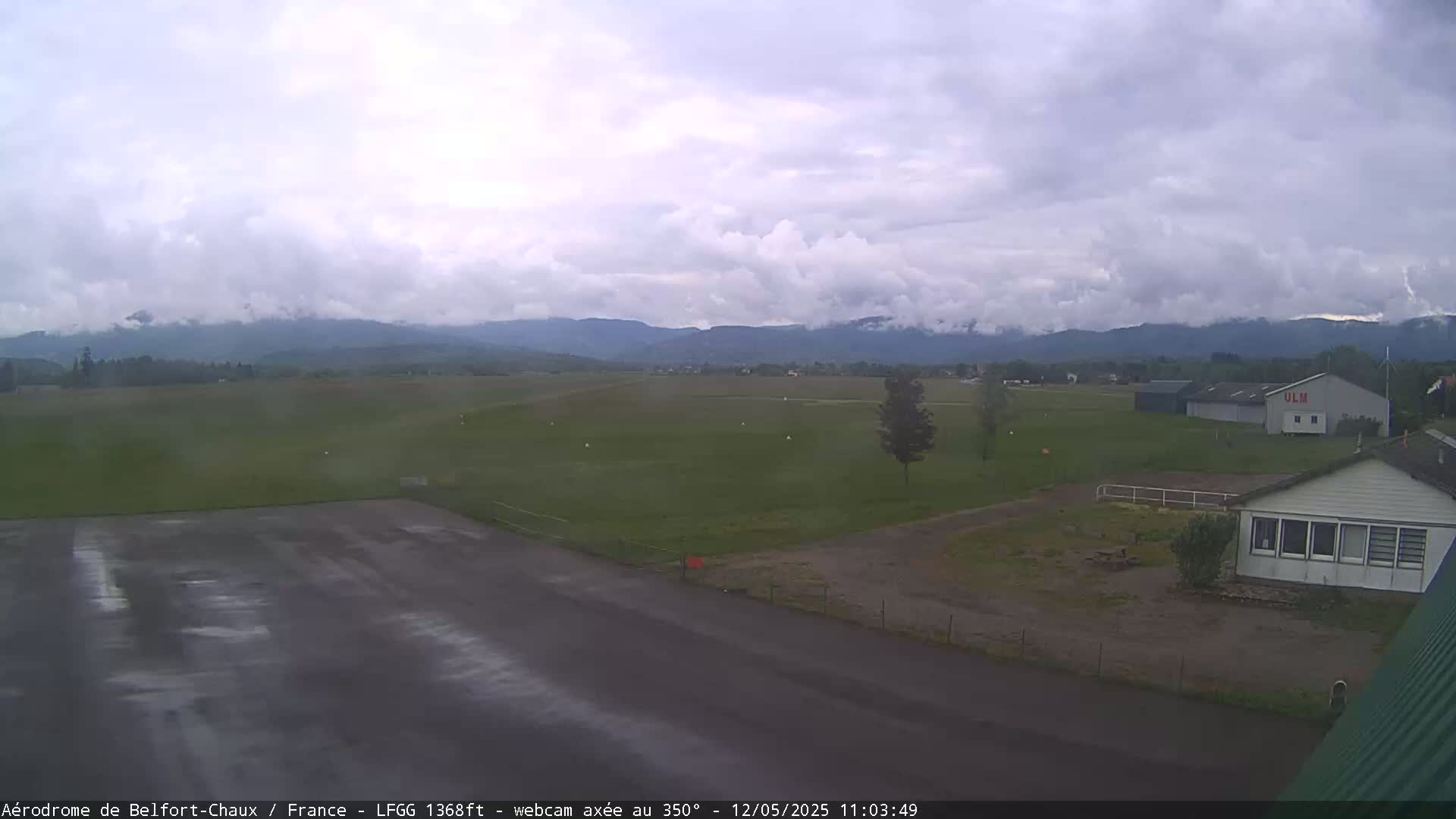 A wet airfield runway sits before a grassy field, a hangar, and a small building, all under a cloudy sky with distant mountains visible.