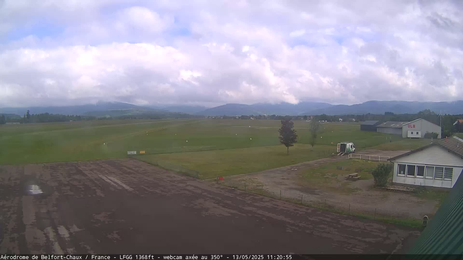 A mostly cloudy day shows a grassy airfield with a runway, a few buildings, and distant mountains.