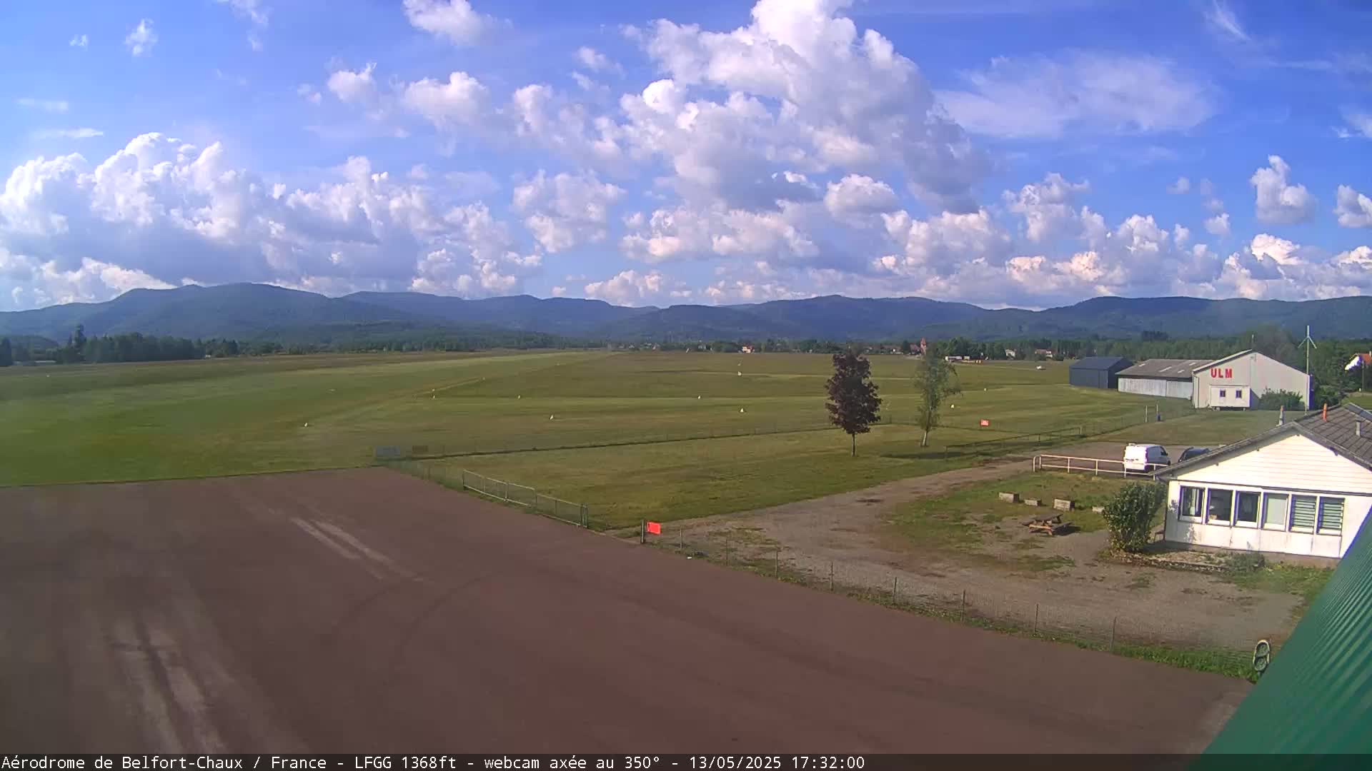 A mostly sunny day with scattered cumulus clouds over a grassy airfield, with a hangar and small building visible, and mountains in the background.