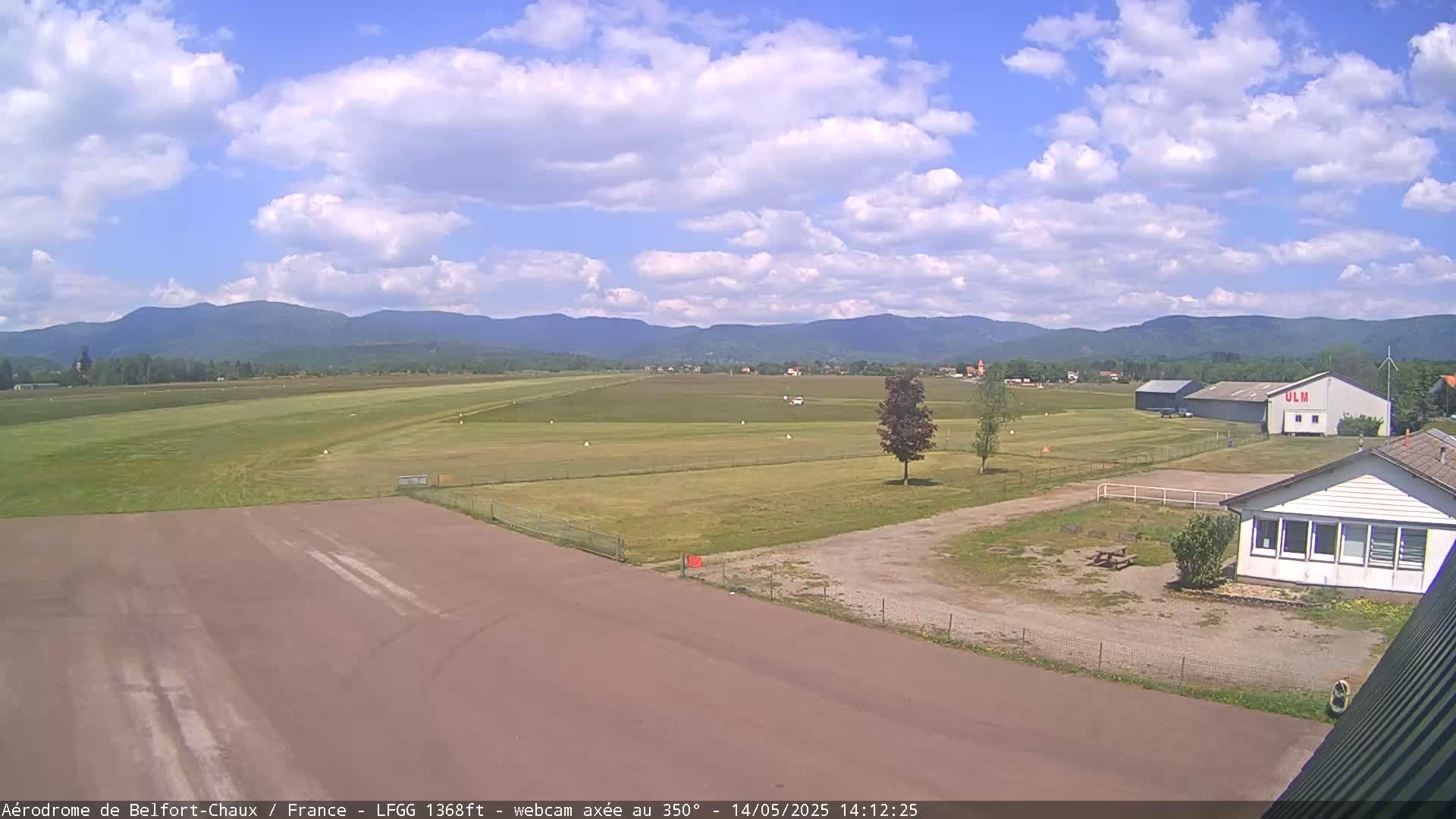 A small airfield with a runway, grassy fields, and a few buildings is seen under a partly cloudy sky with distant mountains in the background.