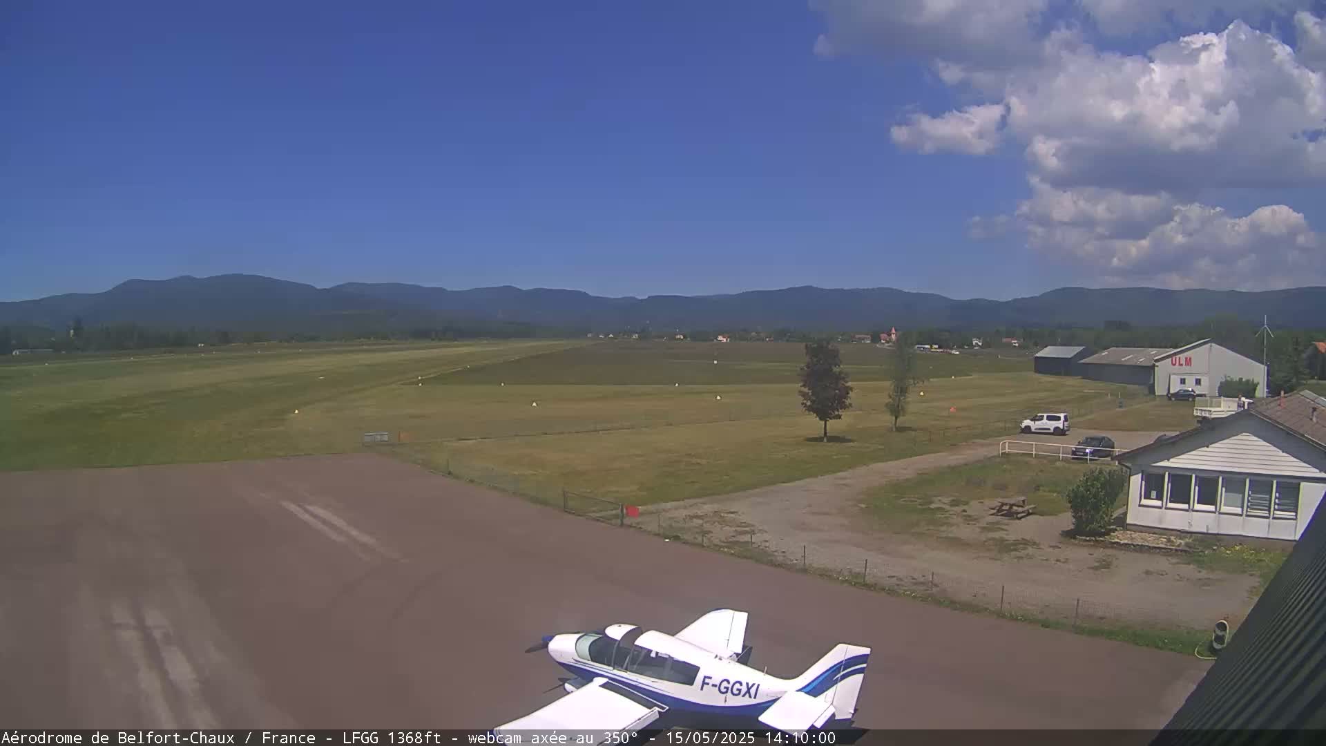 A small airplane sits on a tarmac at an airfield under a partly cloudy sky, with distant mountains in the background.