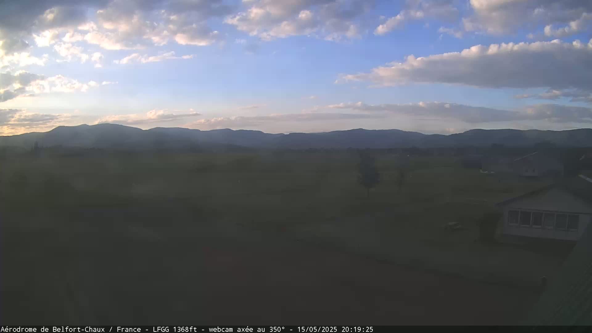 A partly cloudy evening sky over a flat field with distant mountains and a few buildings.