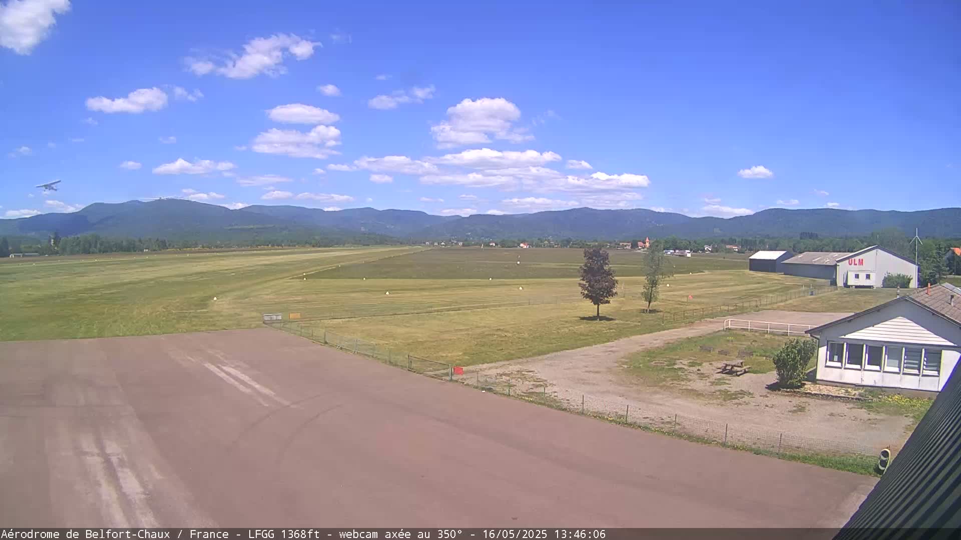 A small airplane flies over a grassy airfield with a few buildings under a mostly sunny sky.