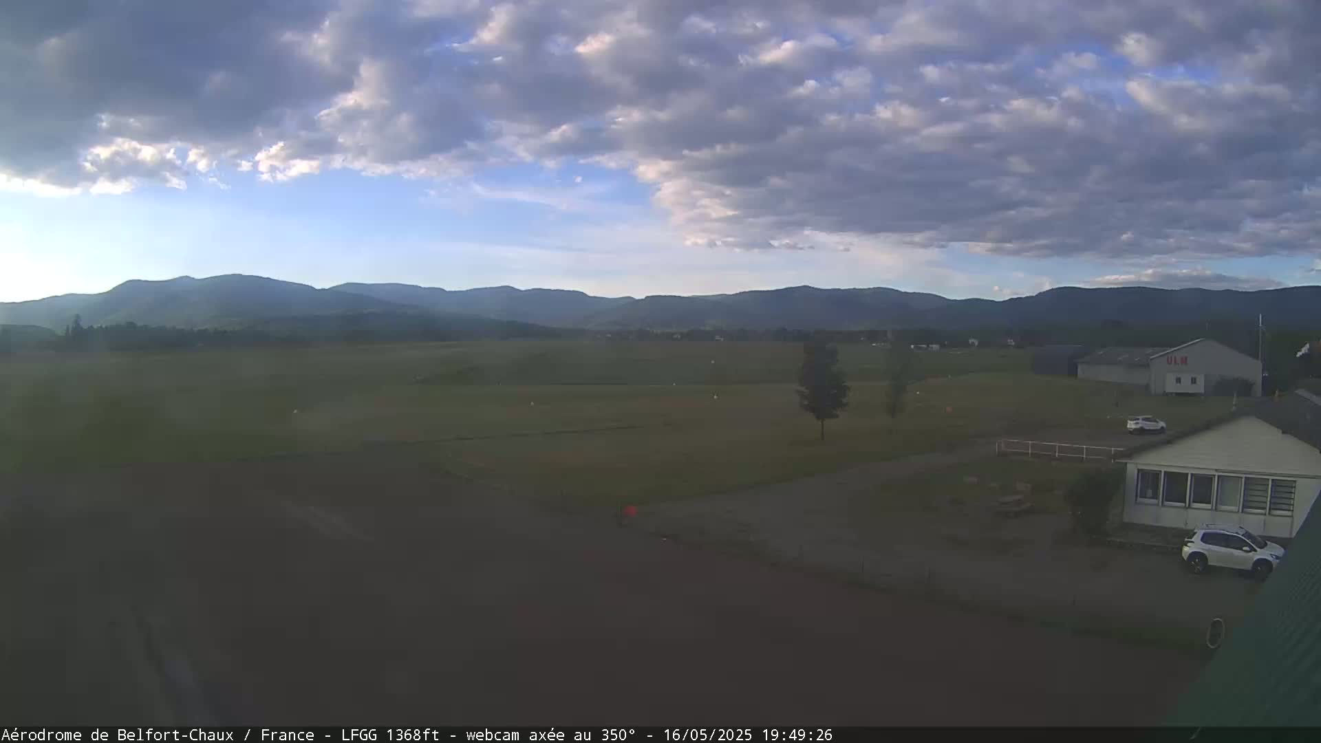 A partly cloudy sky overlooks a flat green field, some buildings, and distant mountains.