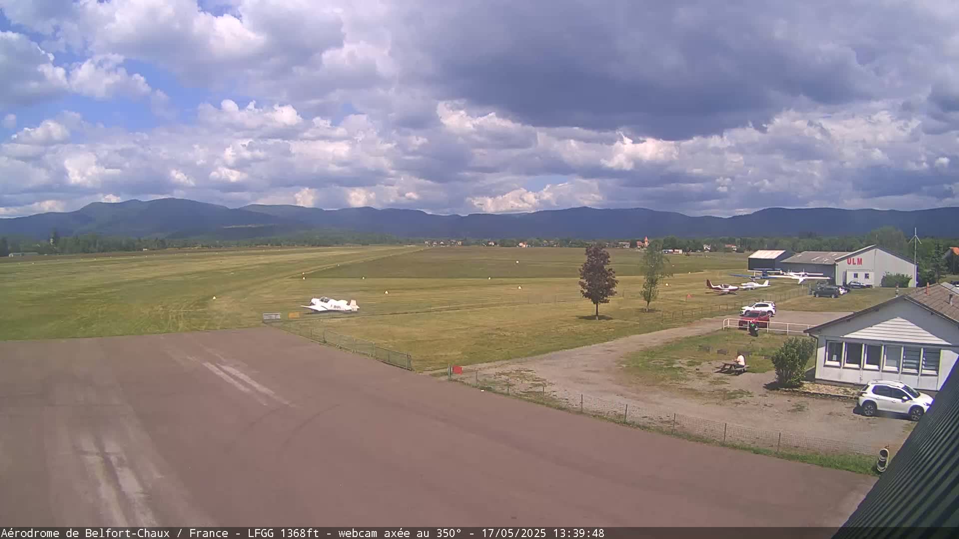 A small airport with several small planes parked near a hangar sits under a partly cloudy sky with mountains in the distance.