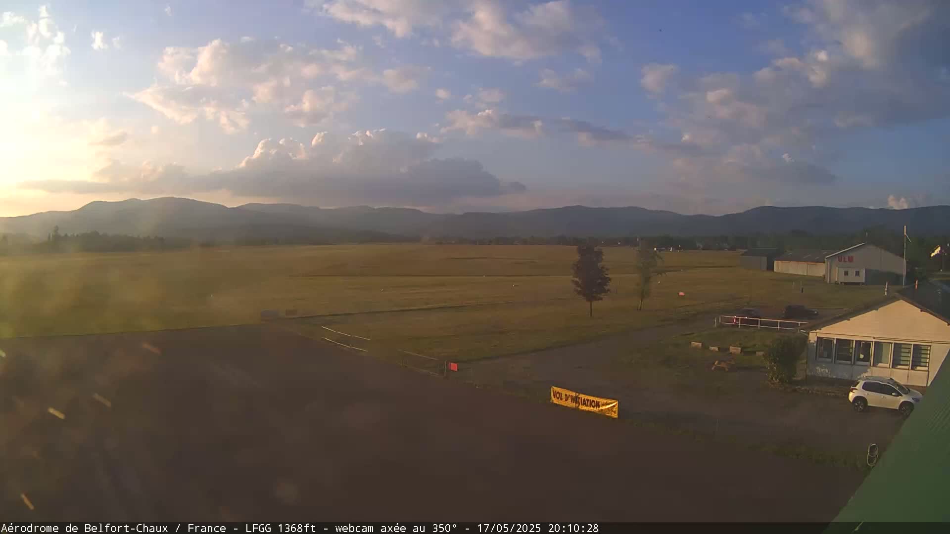 A mostly sunny day shows a flat grassy airfield with a few buildings and a car parked near one, backed by a range of distant mountains under a partly cloudy sky.