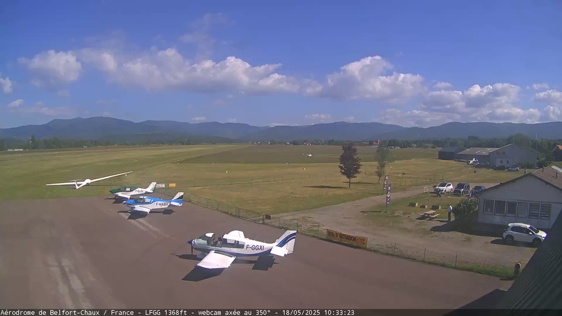 A sunny day at an airfield shows several small airplanes parked on a tarmac with a glider nearby, a hangar in the background, and distant mountains under a partly cloudy sky.