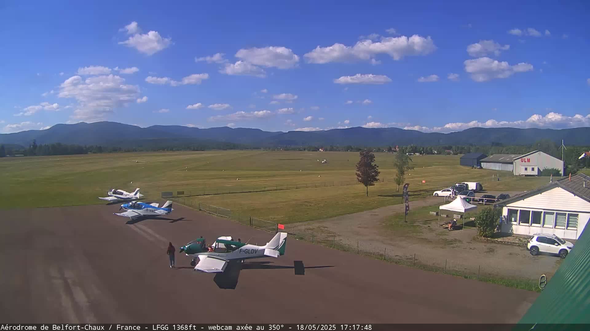 Three small airplanes are parked on a tarmac at an airfield under a partly cloudy blue sky, with mountains visible in the distance.
