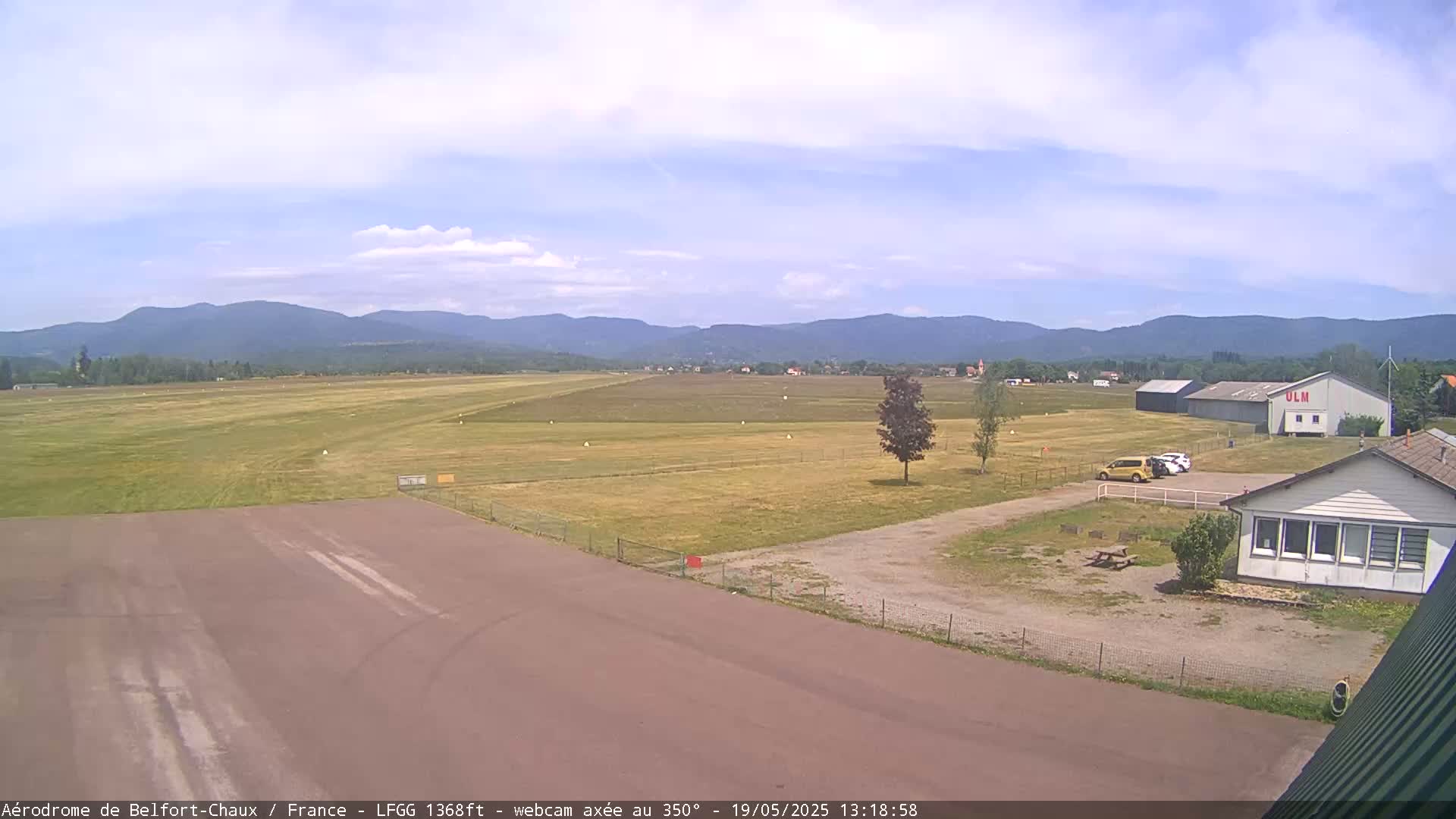 A small airfield with a runway and several buildings is visible under a partly cloudy sky, with mountains in the background.