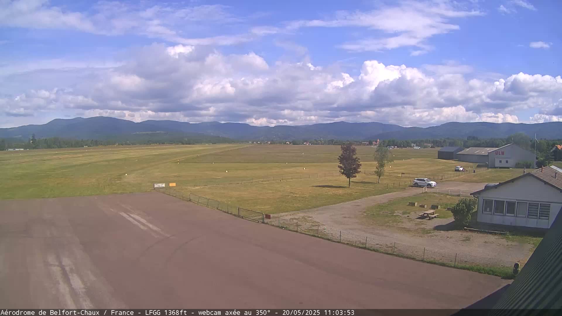 A partly cloudy day at an airfield shows a runway leading to a hangar, a small building, and a few cars parked in a grassy area with mountains in the background.