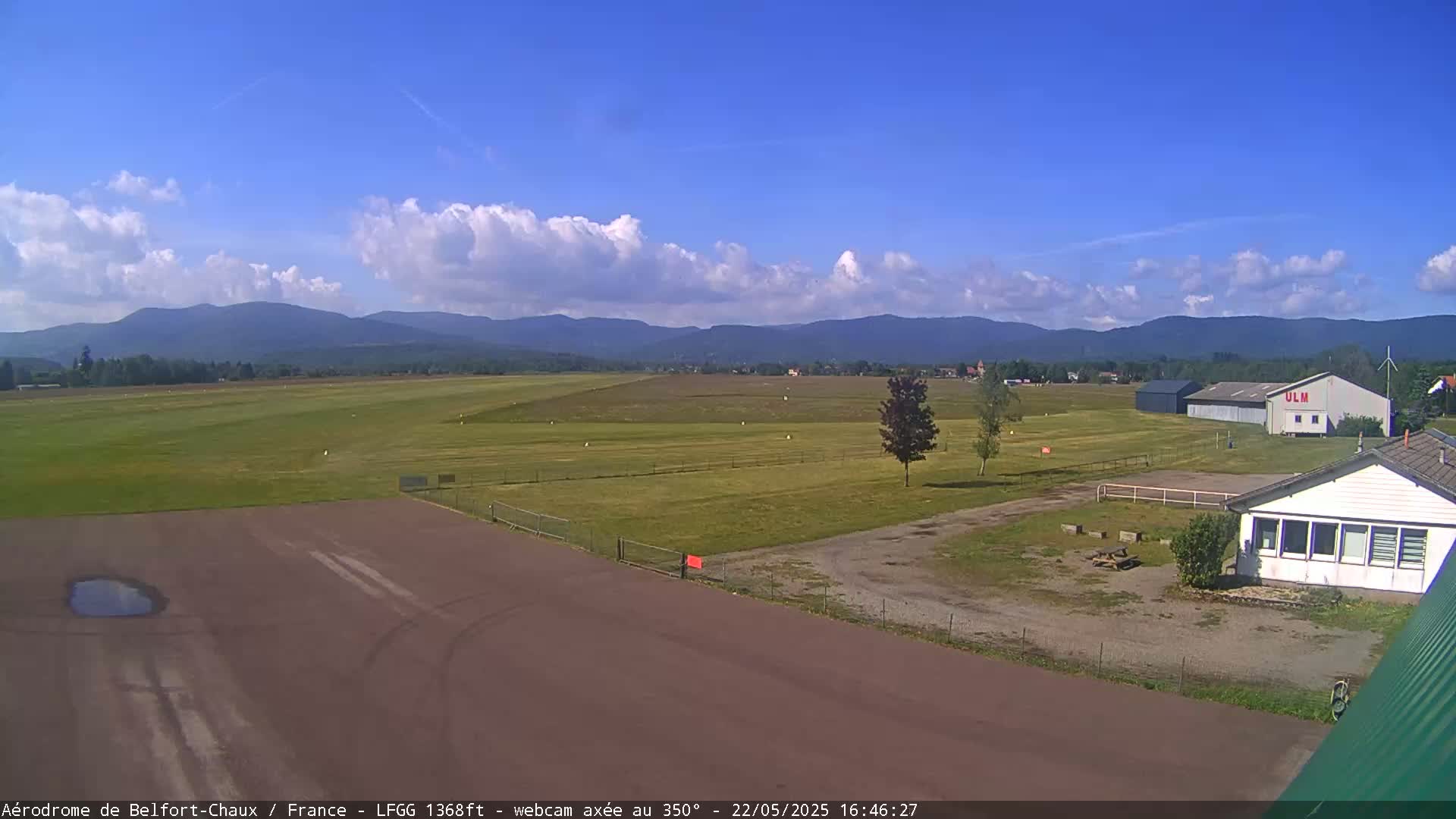 A small airfield with a hangar and small building is visible, set against a backdrop of distant mountains under a partly cloudy blue sky.
