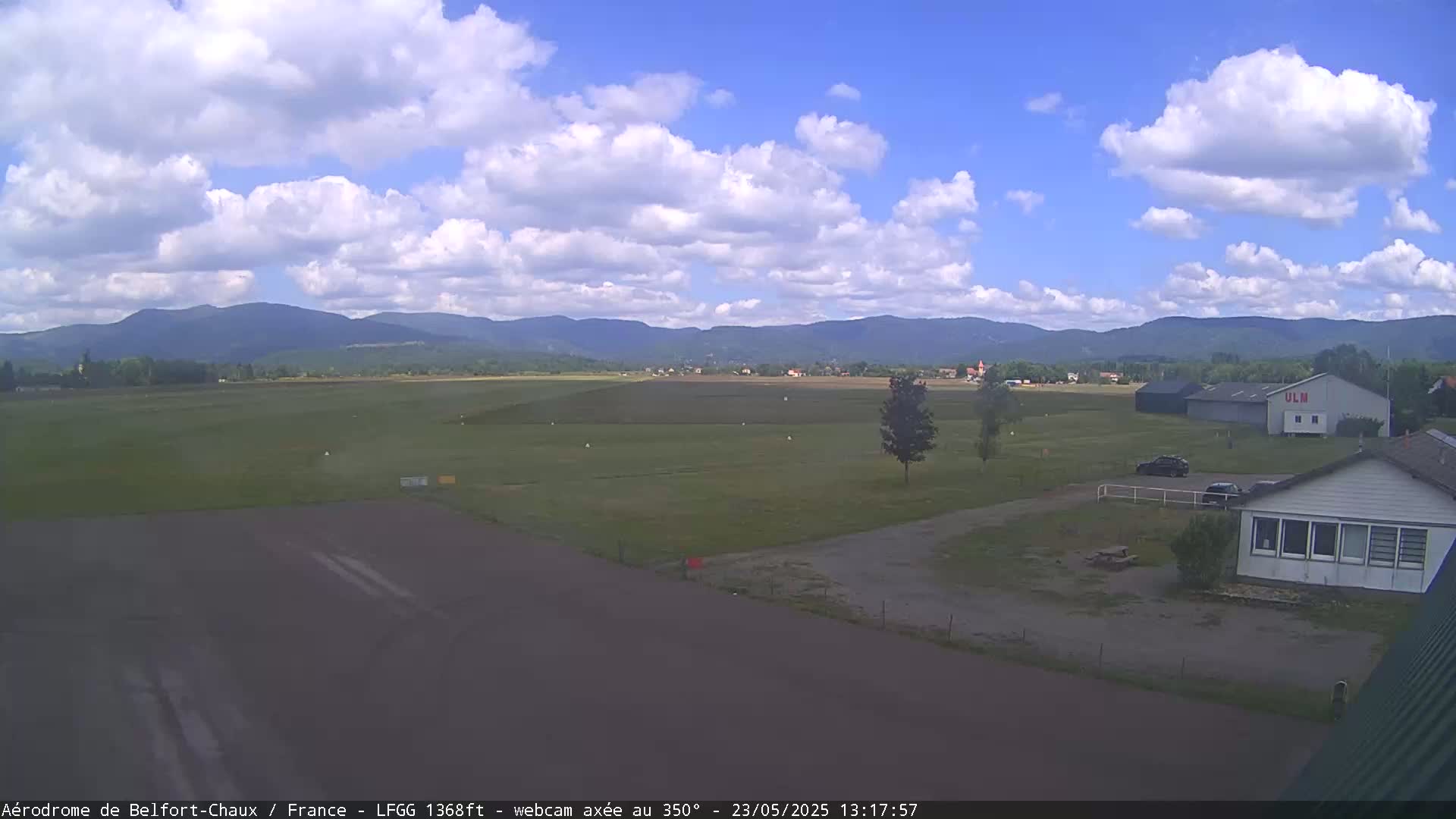 A small airfield with a few buildings and vehicles is visible, set against a backdrop of distant mountains under a partly cloudy sky.