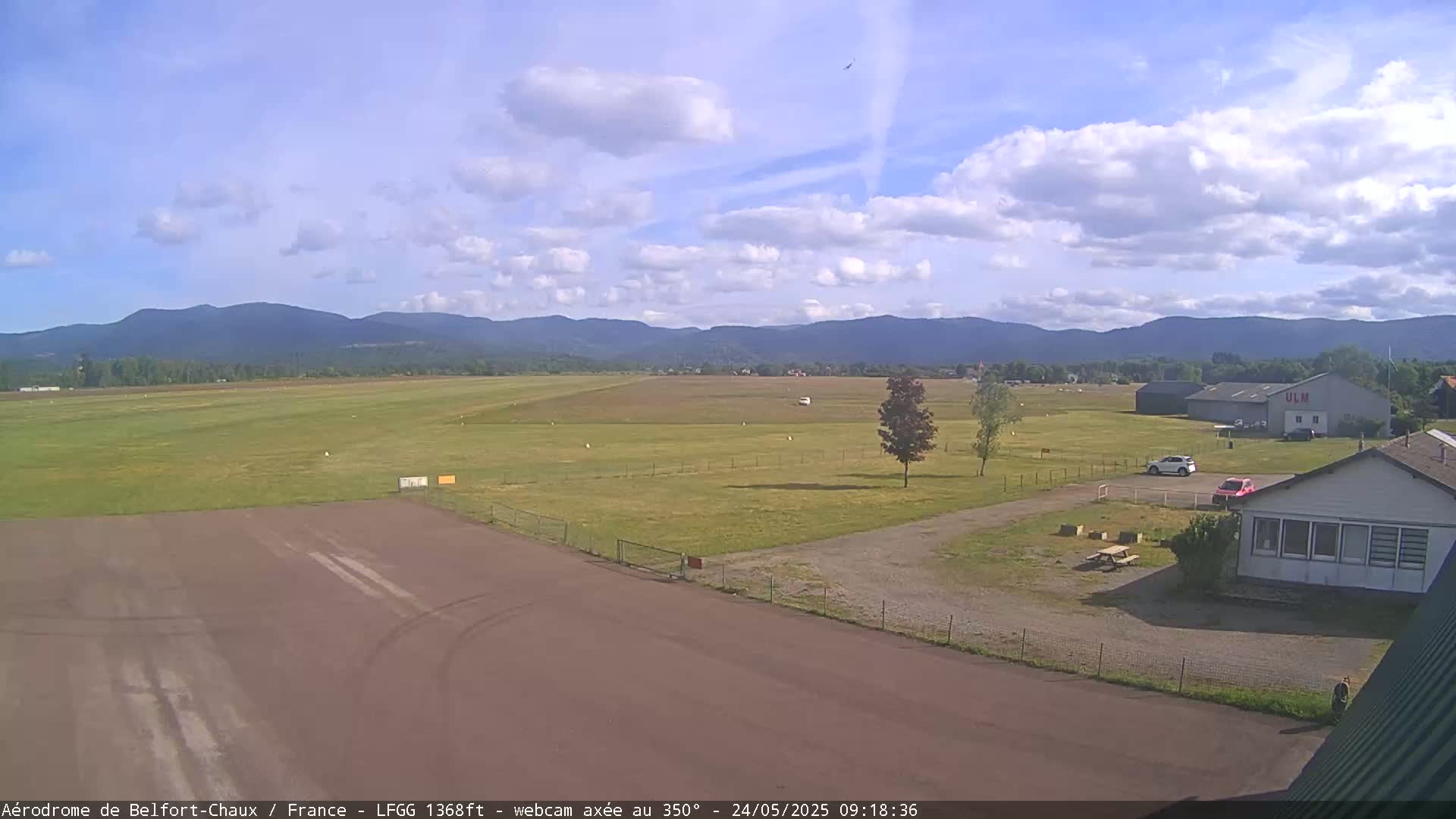 A mostly sunny day shows a small airport with a runway, a few buildings, and some cars, all set against a backdrop of distant mountains.