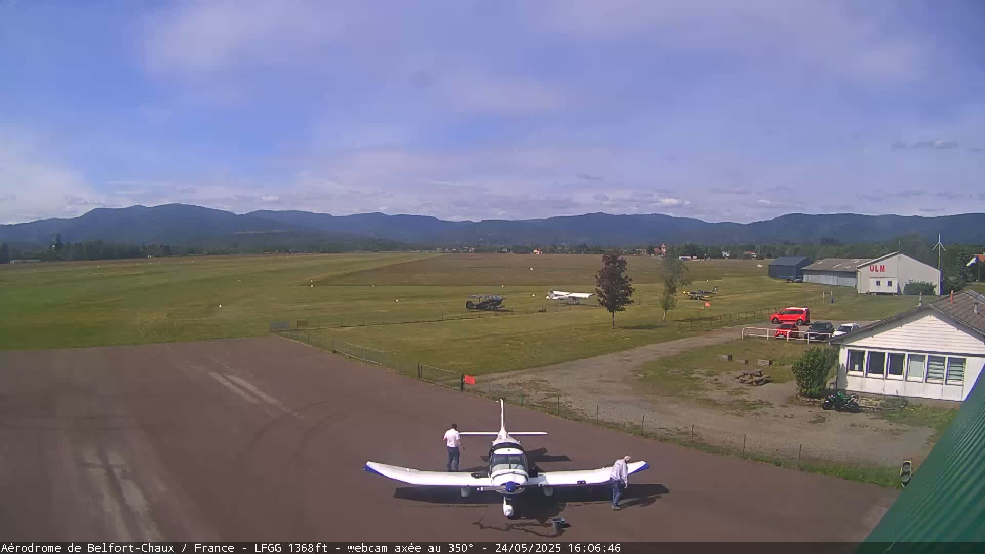 A small airplane sits on a tarmac at an airfield, with two people standing nearby under a partly cloudy sky, and mountains in the distance.