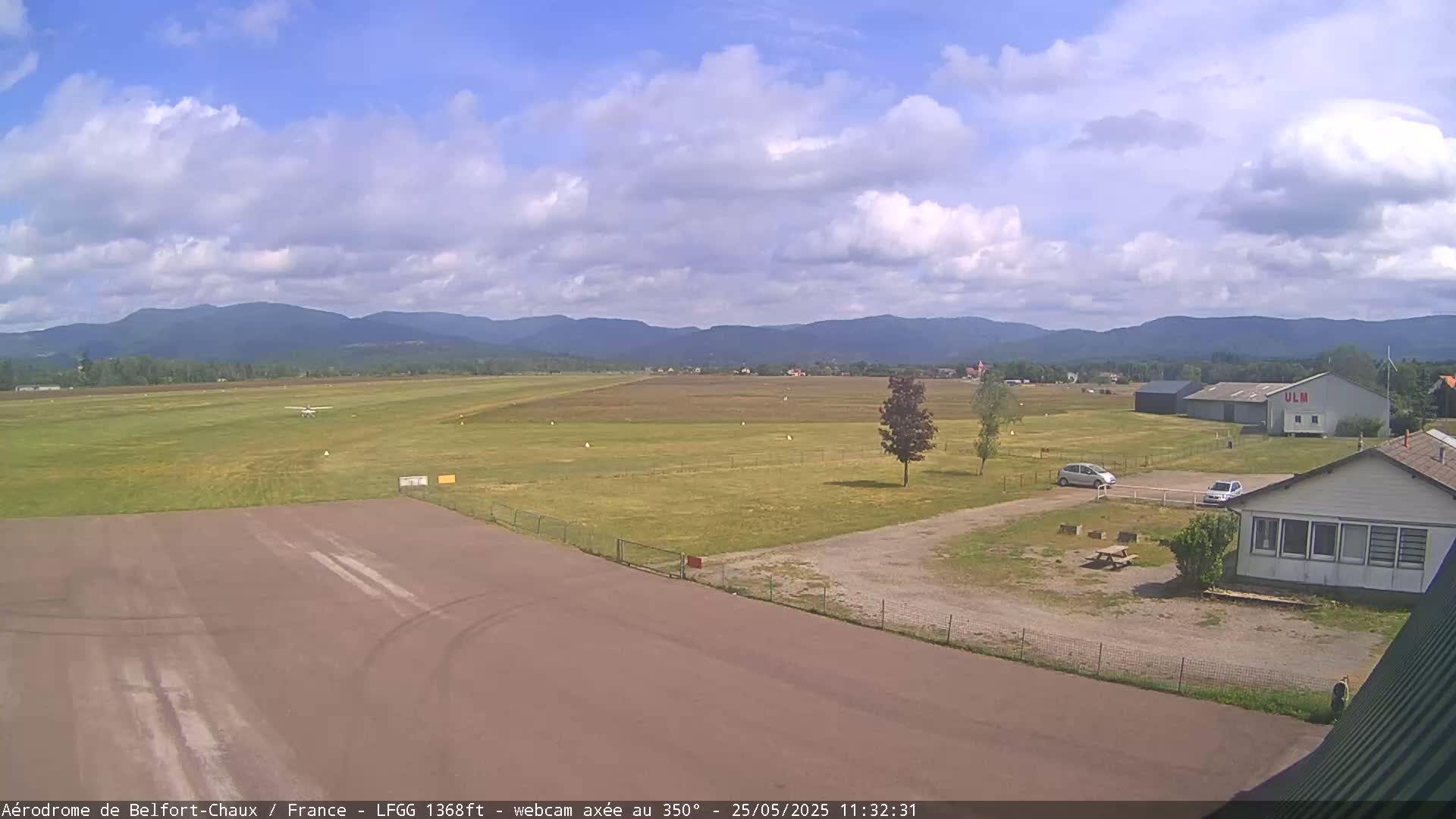A small airfield with a single-engine airplane on the runway is situated before a line of distant mountains under a partly cloudy sky.